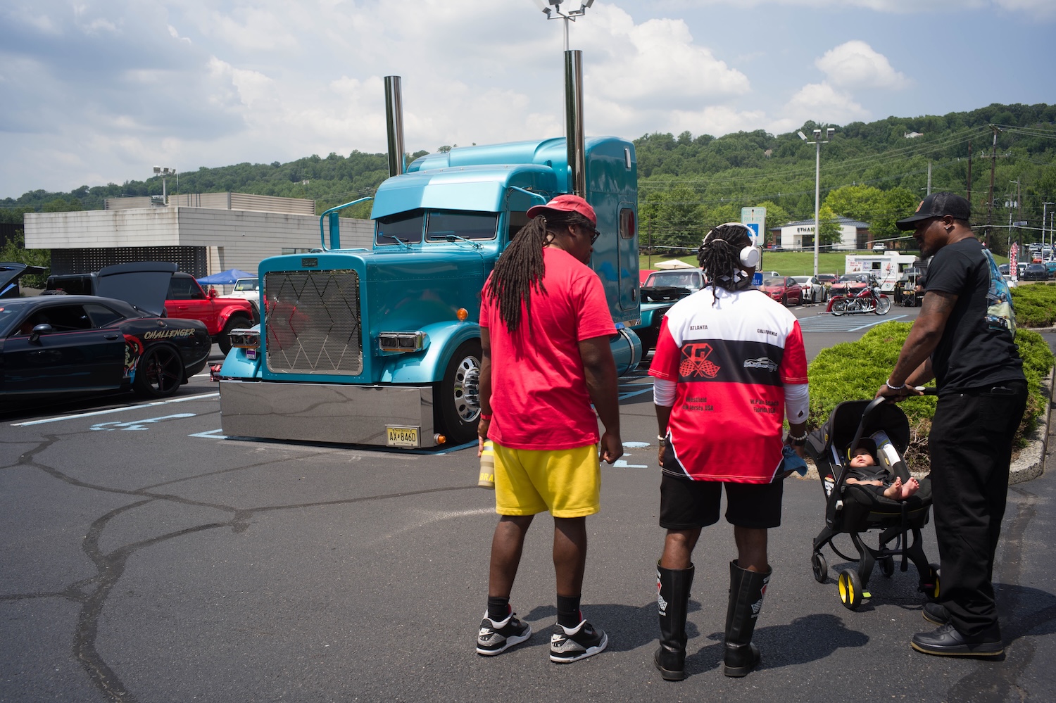 Man in red shirt with woman in racing jersey and a man with a baby in a stroller, standing in front of a teal big rig truck.