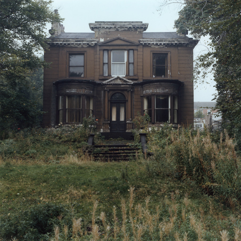 A dilapidated brown house with overgrown grass and weeds surrounding it, featuring large windows and a staircase leading to the entrance.