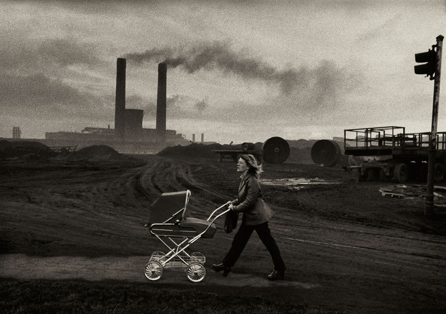 A woman pushing a stroller walks along a dirt path, with smokestacks and industrial machinery in the background, under a cloudy sky.