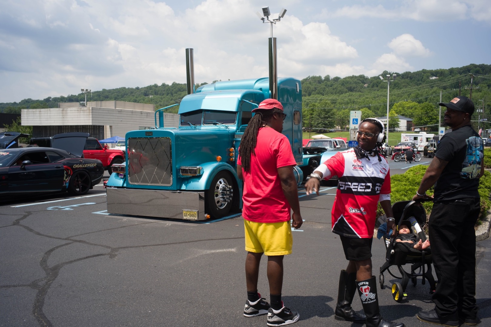 Man in red shirt with woman in racing jersey and a man with a baby in a stroller, standing in front of a teal big rig truck.