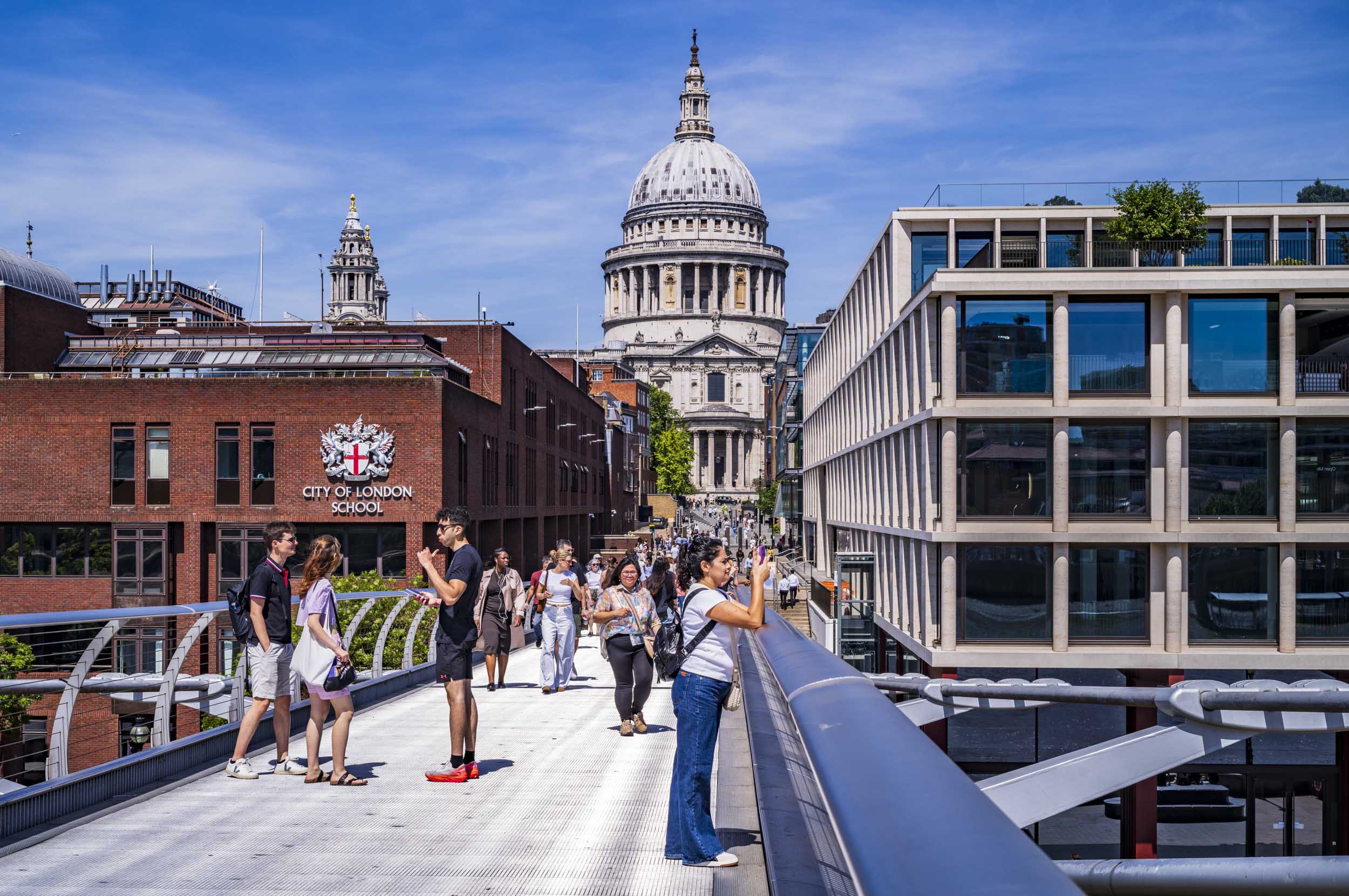 The dome of St Paul's Cathedral rising above surrounding buildings, seen from the Millennium Bridge in London, with pedestrians walking and taking photos on the bridge and the City of London School building to the left.