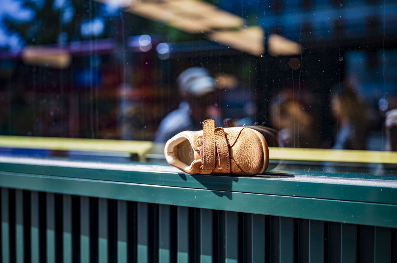 A small tan leather child's shoe with a buckle strap lying on its side on a green railing, with blurred figures and a market stall visible through glass in the background.