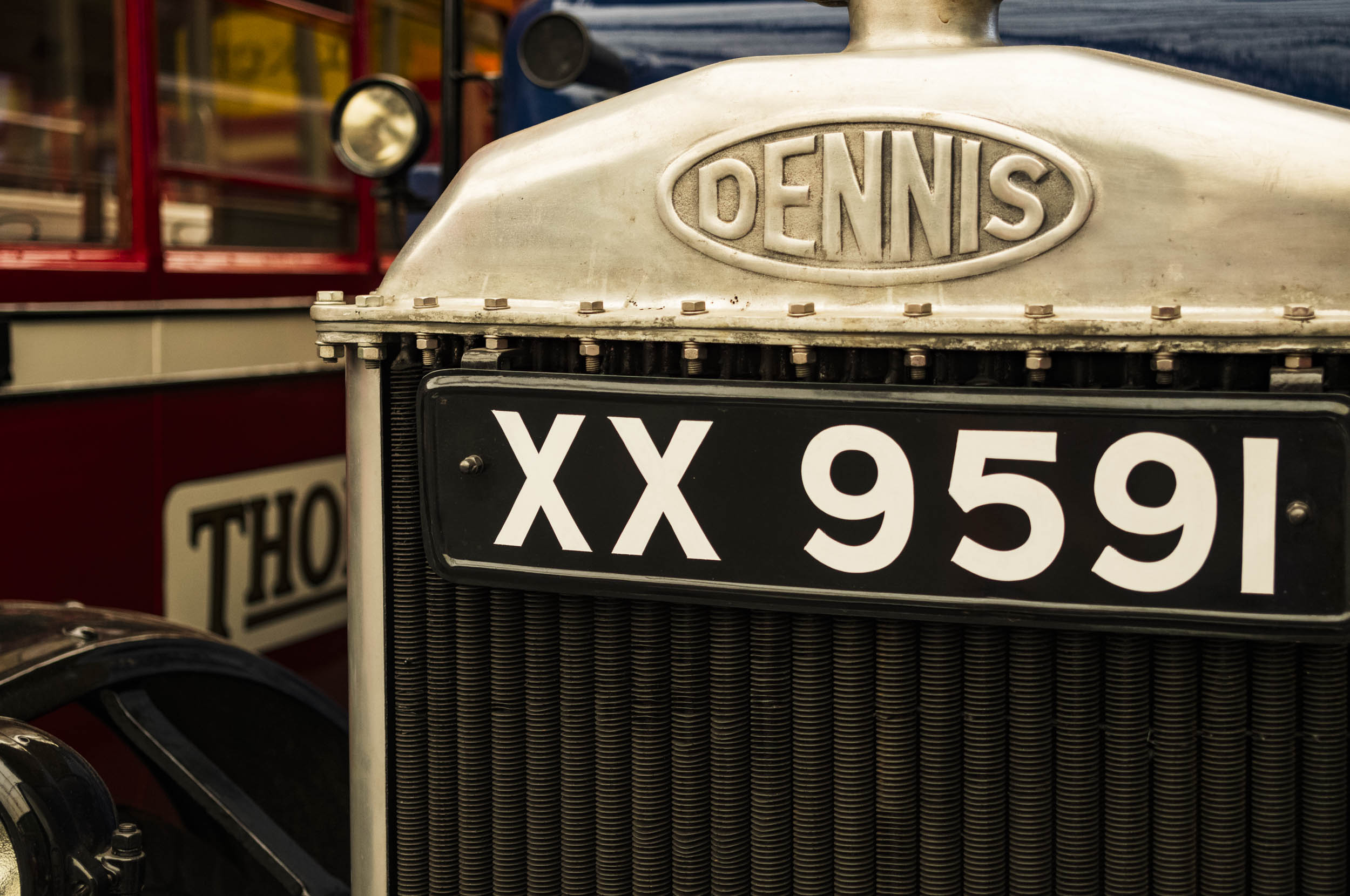 Dennis radiator badge and headlamp on a vintage preserved bus, with a black number plate reading "XX 9591" below the grille, another red double-decker bus visible out of focus in the background.