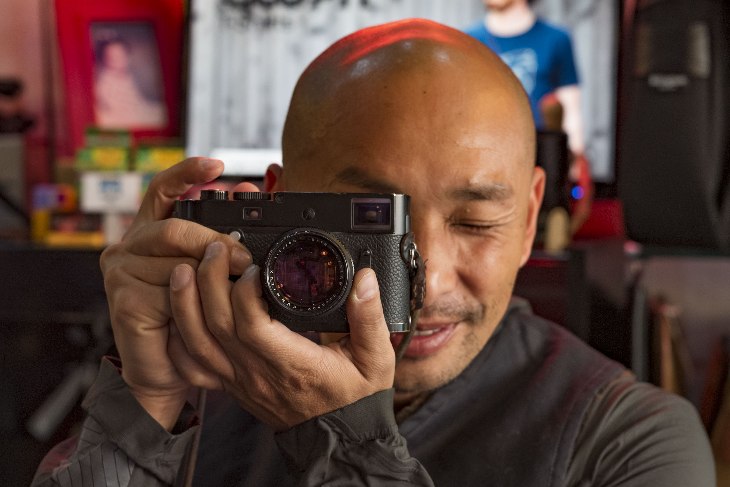 Close-up of a bald man smiling while looking down through the viewfinder of a black rangefinder camera with a large lens, in a colorful indoor setting with blurred figures in the background.