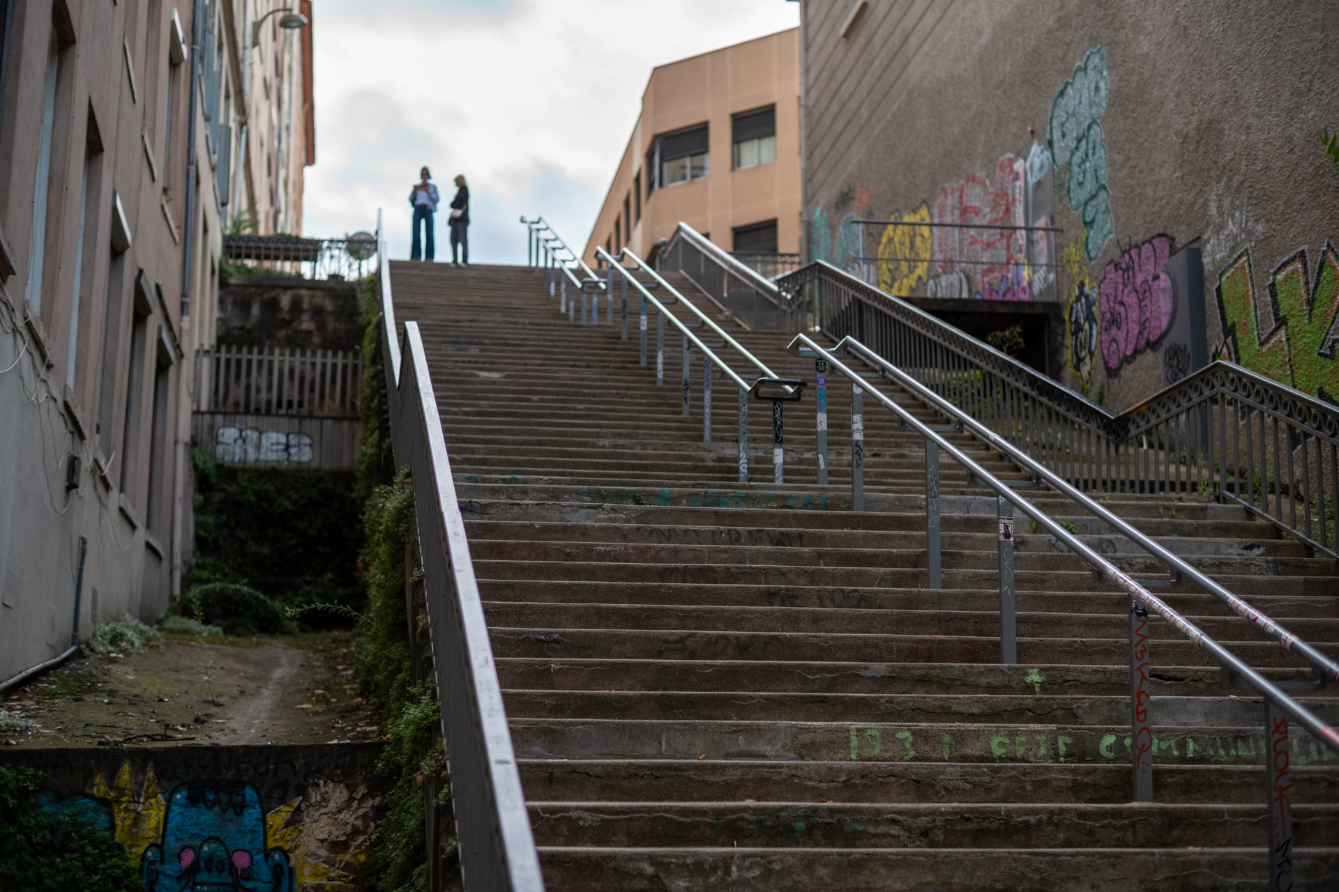 Sample shot with Thypoch Simera 50/1.4: a graffiti-lined staircase between buildings, with two people talking at the top, Lyon, France.