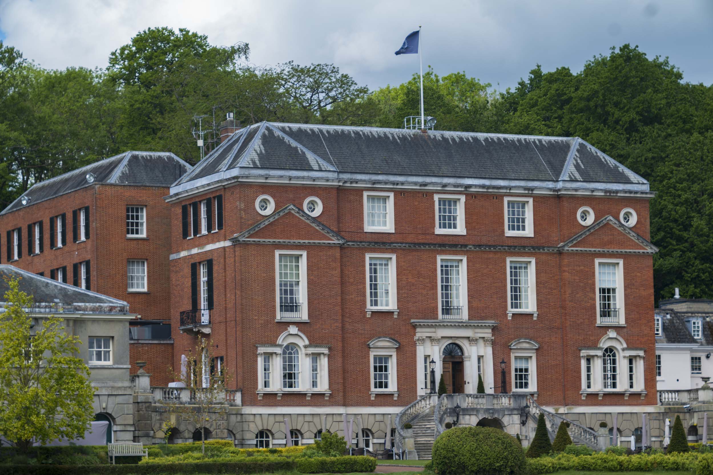 Grand red-brick Georgian-style mansion with white-trimmed windows, decorative pediments, a flagpole flying a flag on the roof, and a curved stone staircase leading up to the entrance, surrounded by trees and manicured hedges. RAC Country Club, Epsom.