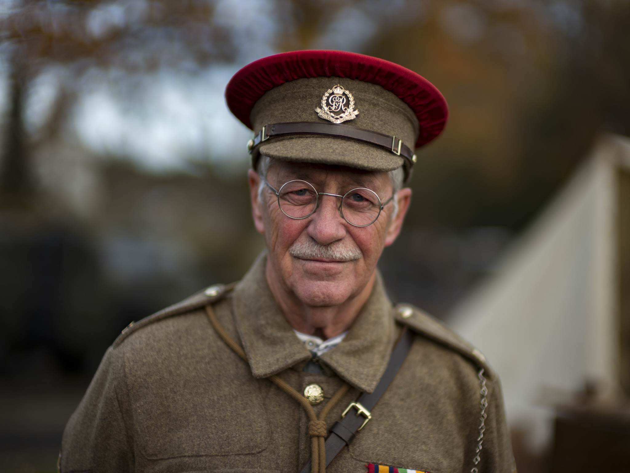 Portrait of an elderly man in vintage military uniform, wearing round wire-rimmed glasses and a khaki peaked cap with a red band and Military Police badge, with a white mustache, standing outdoors against a blurred autumn background.