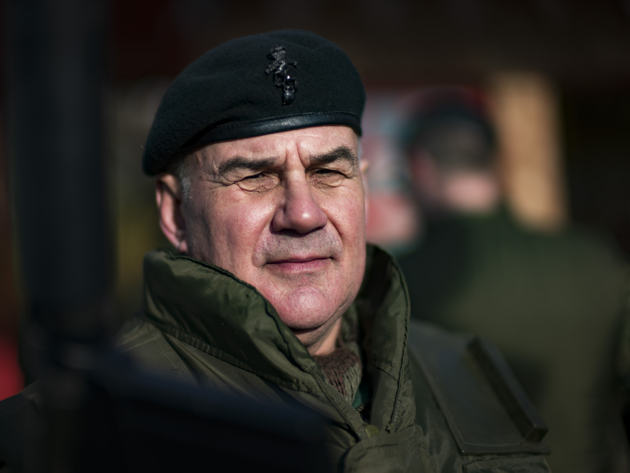 Close-up portrait of an older man wearing a dark green military beret with a badge and a green jacket with a raised collar, looking off to the side with a slight smile against a blurred dark background.