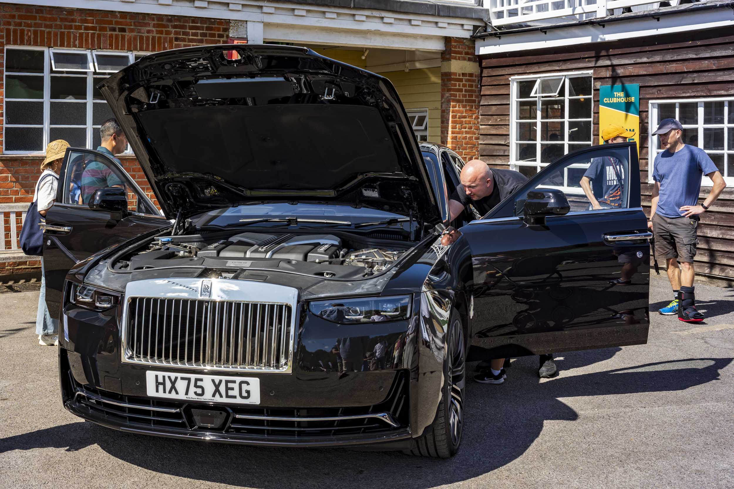A man leans into the open bonnet of a modern black Rolls-Royce saloon to inspect the engine, with onlookers gathered around outside a clubhouse building.