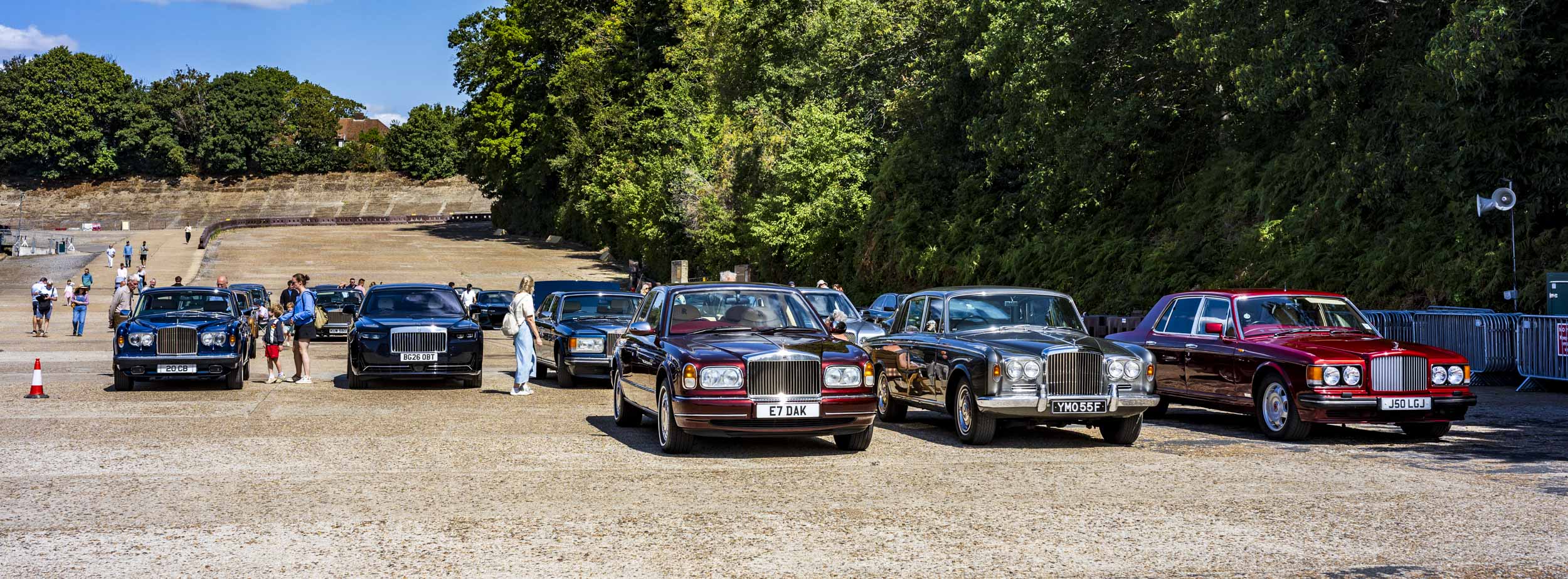 A line-up of classic Rolls-Royce and Bentley cars parked on a gravel area with visitors walking among them, a banked concrete race track visible behind.