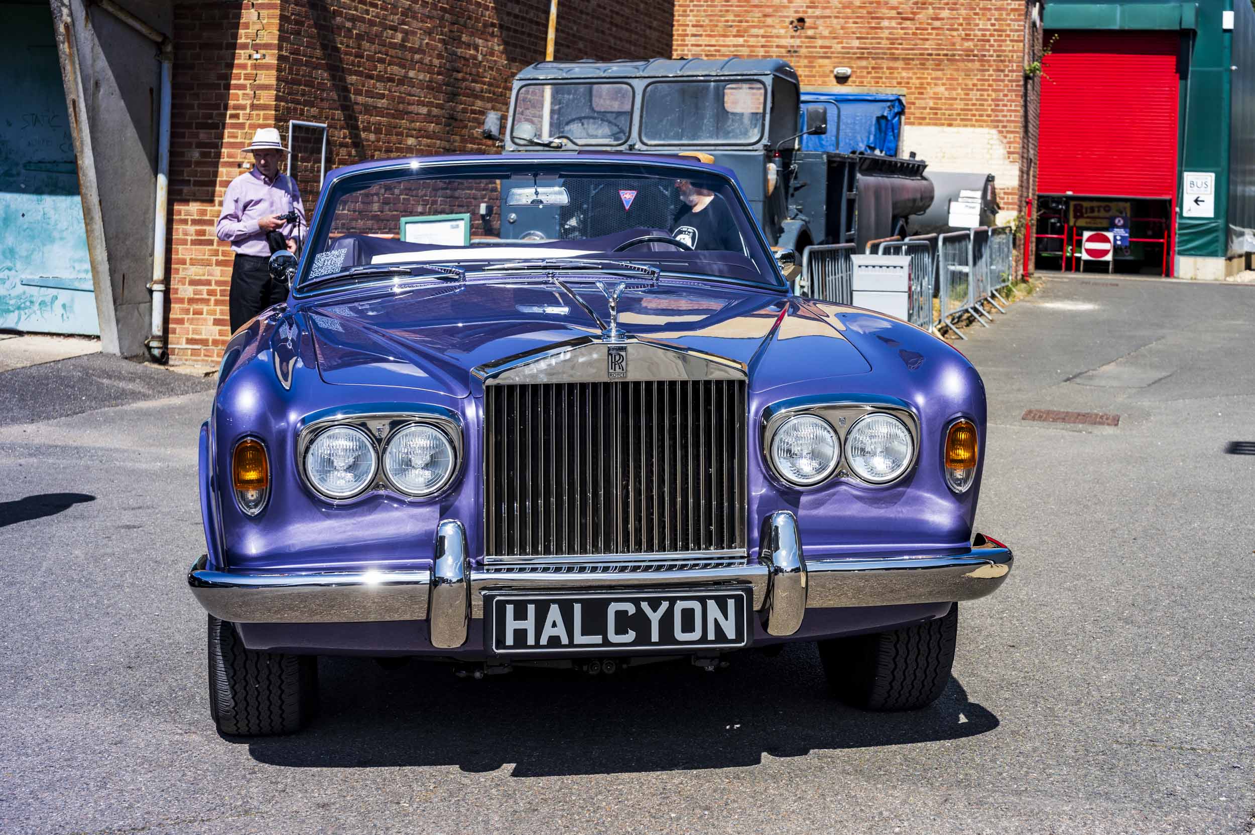 Front view of a purple convertible Rolls-Royce Corniche with a "HALCYON" number plate, parked beside a brick building and an old truck.