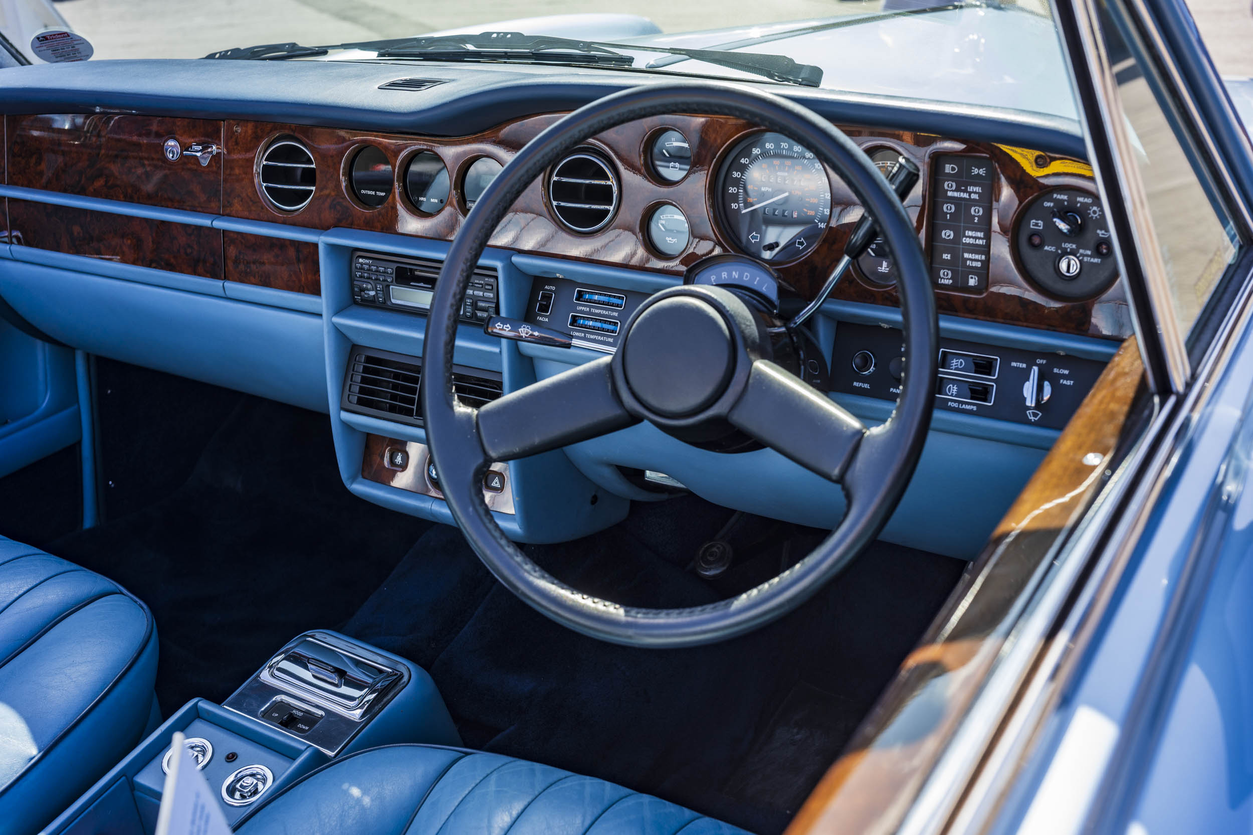 Interior view of a classic Rolls-Royce dashboard with blue leather trim, burr walnut veneer, steering wheel and instrument cluster.