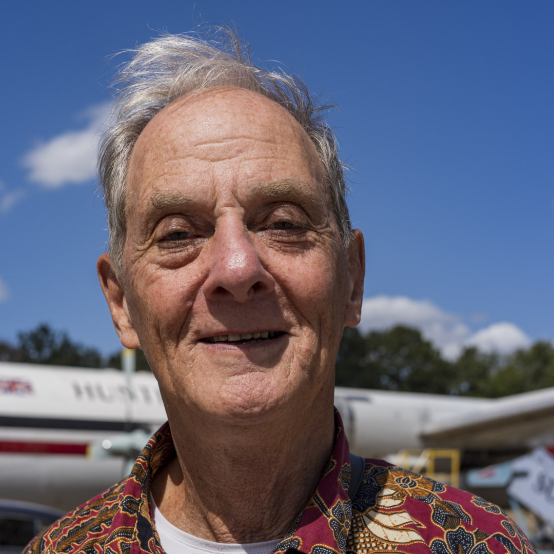 Portrait of a smiling elderly man with white hair wearing a patterned red shirt, standing in front of a vintage aircraft.