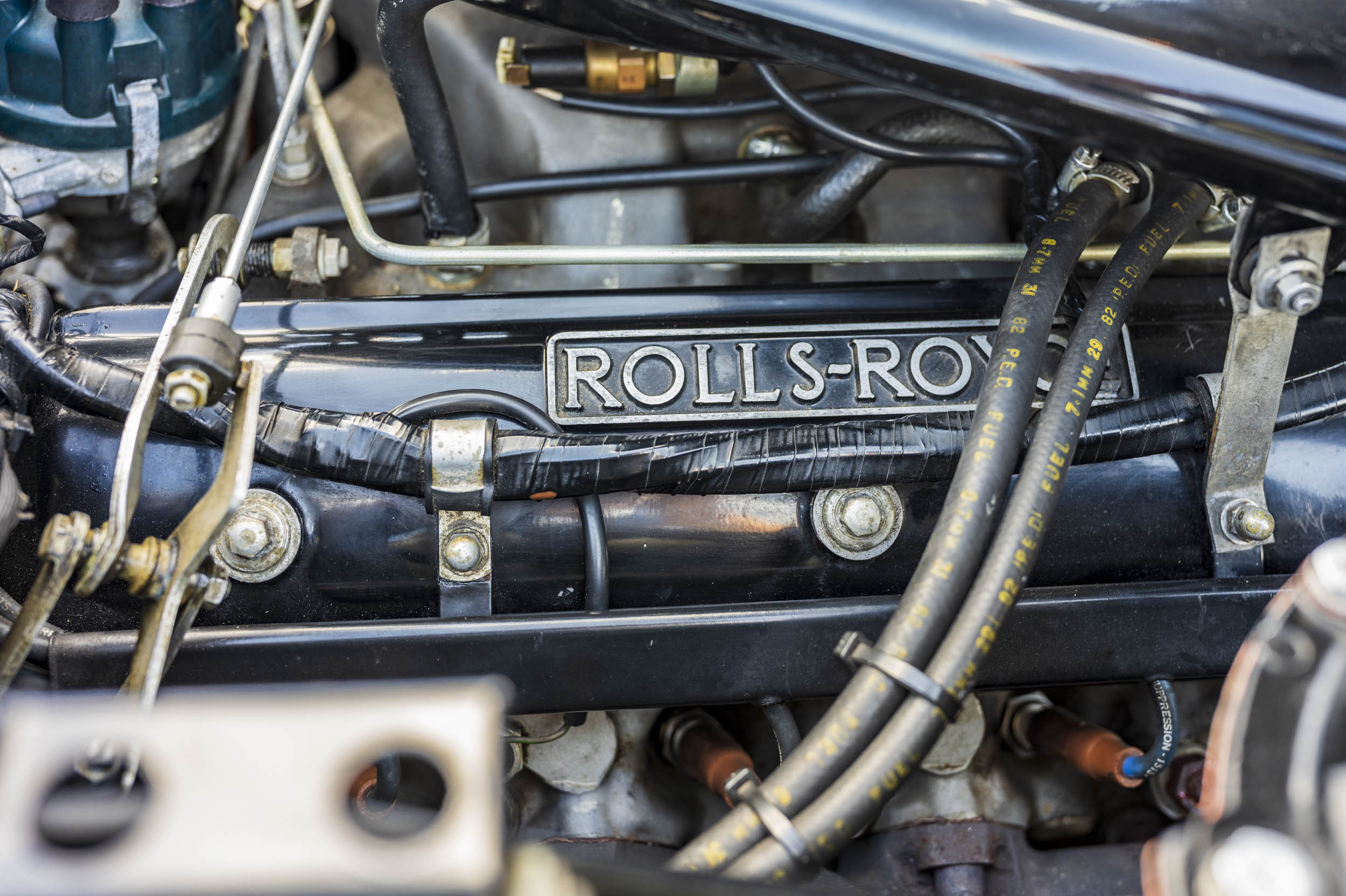 Close-up of an engine bay with a "ROLLS-ROYCE" badge on the rocker cover, surrounded by fuel lines and hoses.