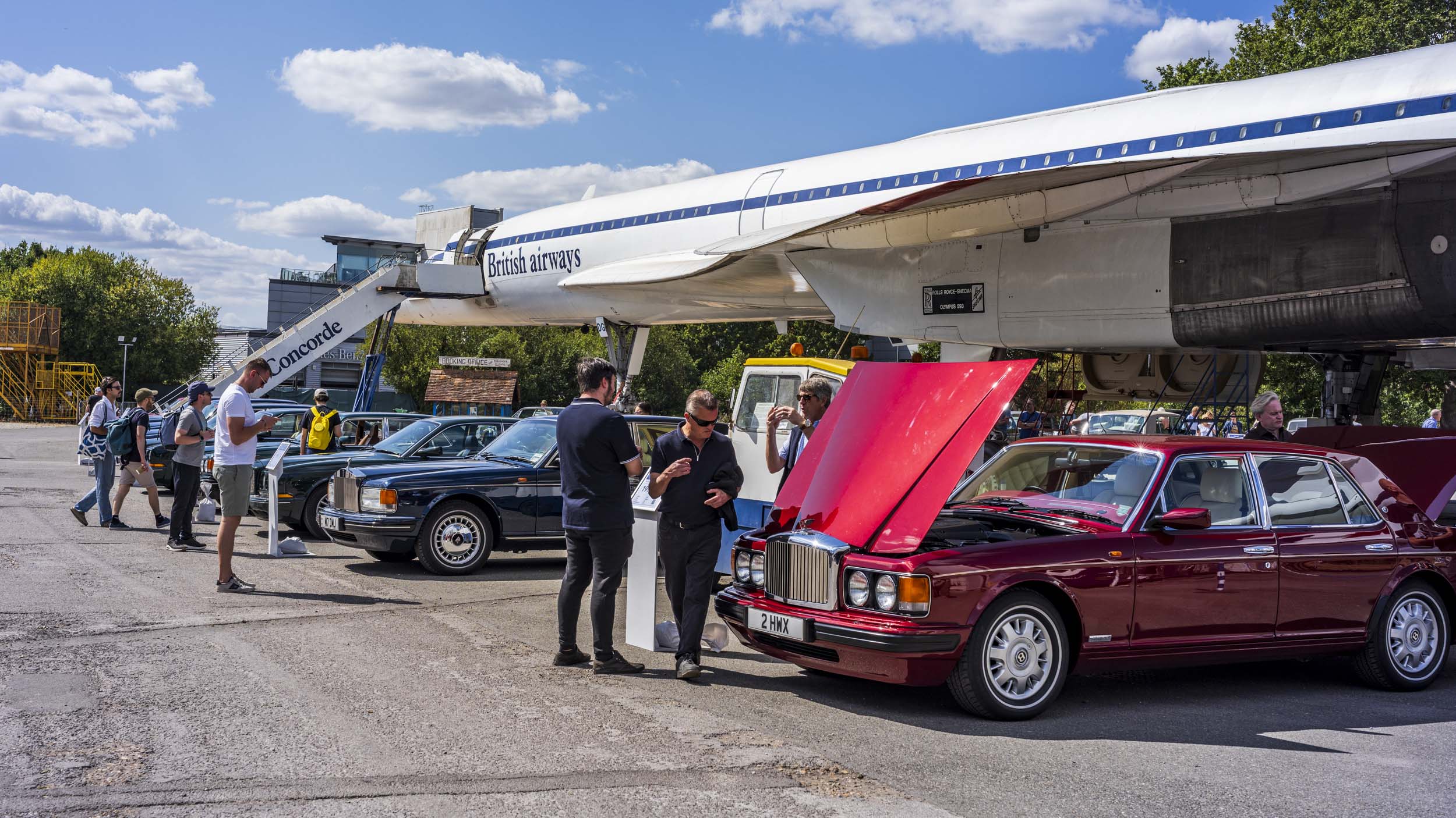 A dark red Bentley Turbo R with its bonnet open, parked beneath the nose of Concorde at a museum, with people examining the car.