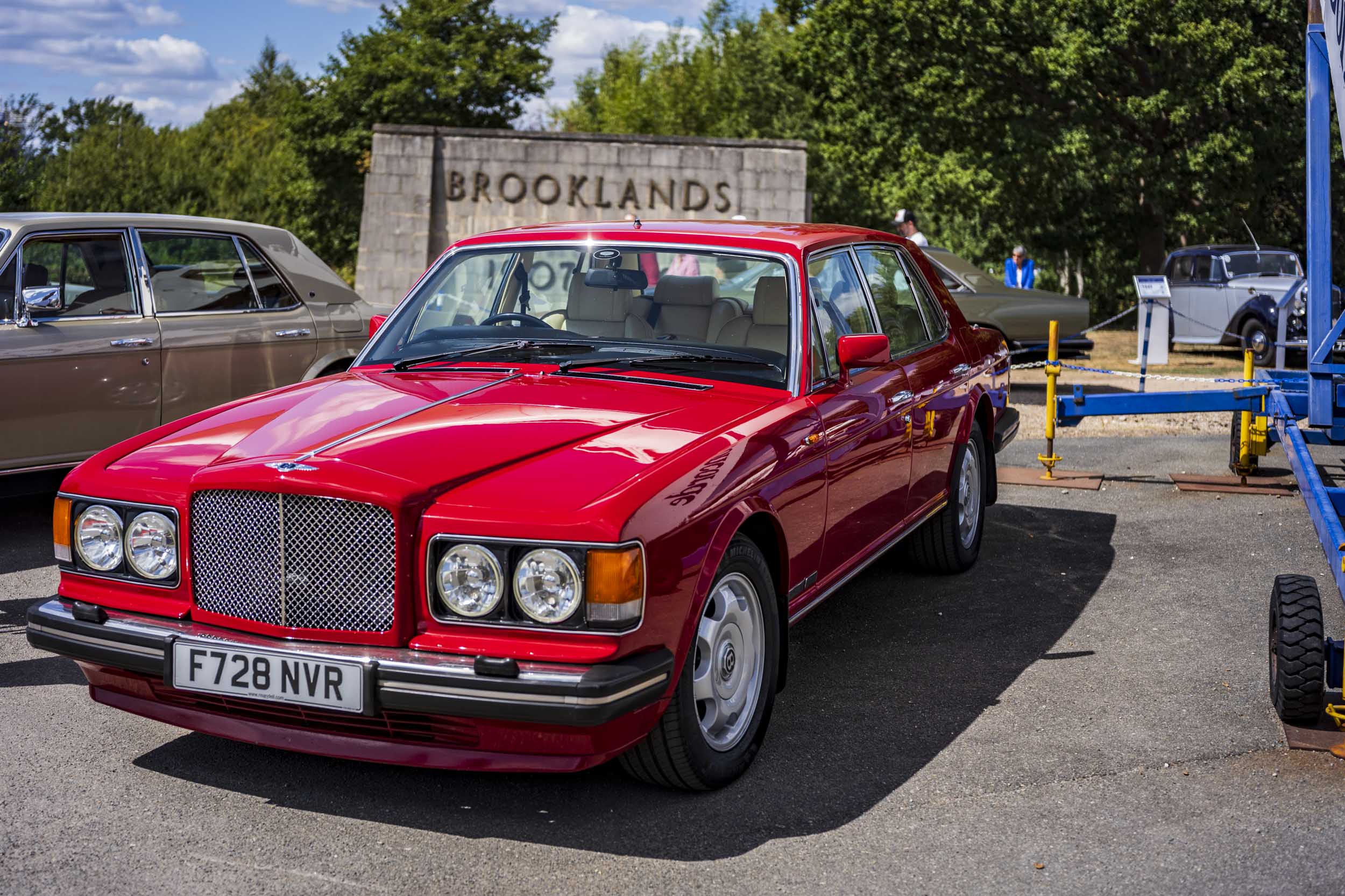 A red Bentley Turbo R saloon car on display at a classic car show, parked in front of the Brooklands sign.