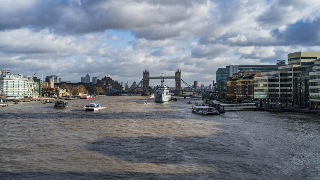 View down the River Thames toward Tower Bridge, with HMS Belfast moored in the foreground, riverboats crossing the choppy water, and buildings lining both banks under a partly cloudy sky.
