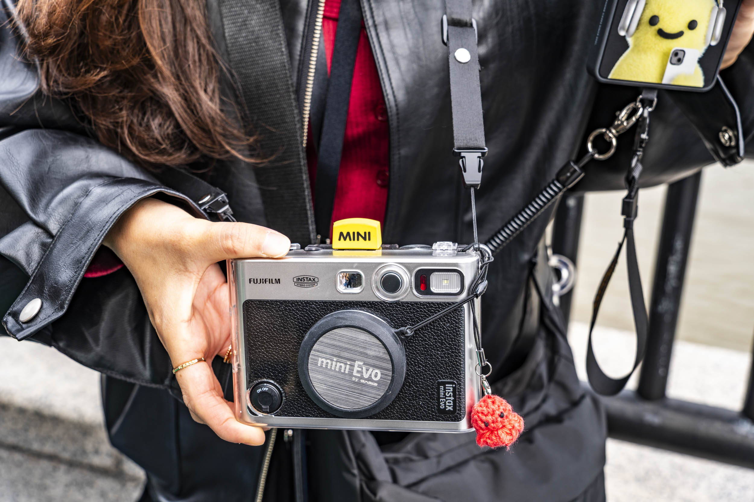 Close-up of a woman's hand holding a Fujifilm Instax Mini Evo camera, with a red crochet charm attached to the strap.