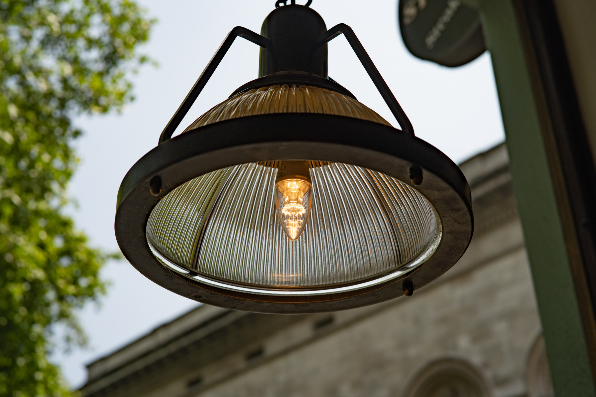 Vintage industrial pendant light fixture with a ribbed glass dome shade and visible glowing filament bulb, hanging against a pale sky with green tree foliage in the upper left and a building facade in the background.