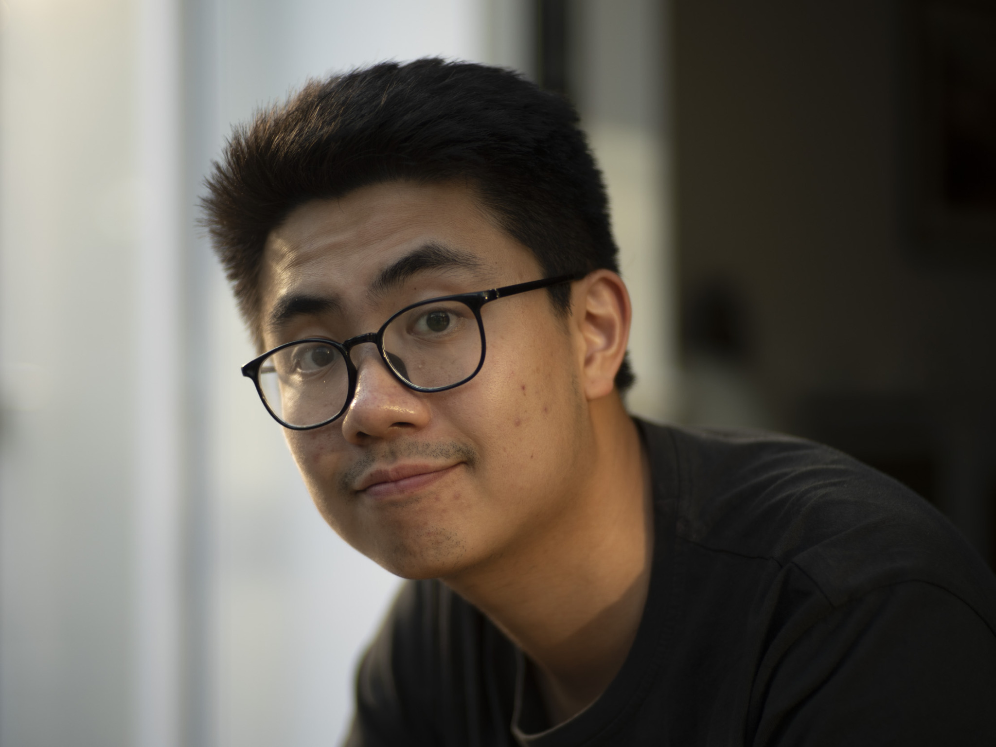 Close-up portrait of a young man with short dark hair, wearing black-framed glasses and a dark shirt, looking at the camera with a slight smile against a blurred indoor background.
