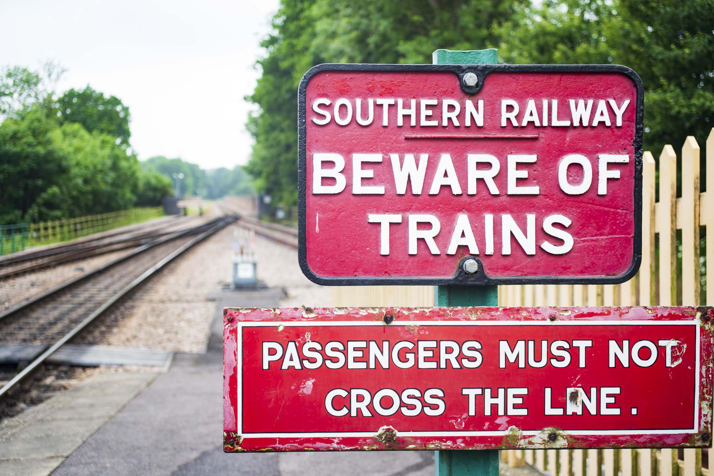 Southern Railway "Beware of Trains" sign with "Passengers Must Not Cross the Line" sign below, mounted on a green post at a railway crossing, with tracks and a wooden picket fence visible in the background.