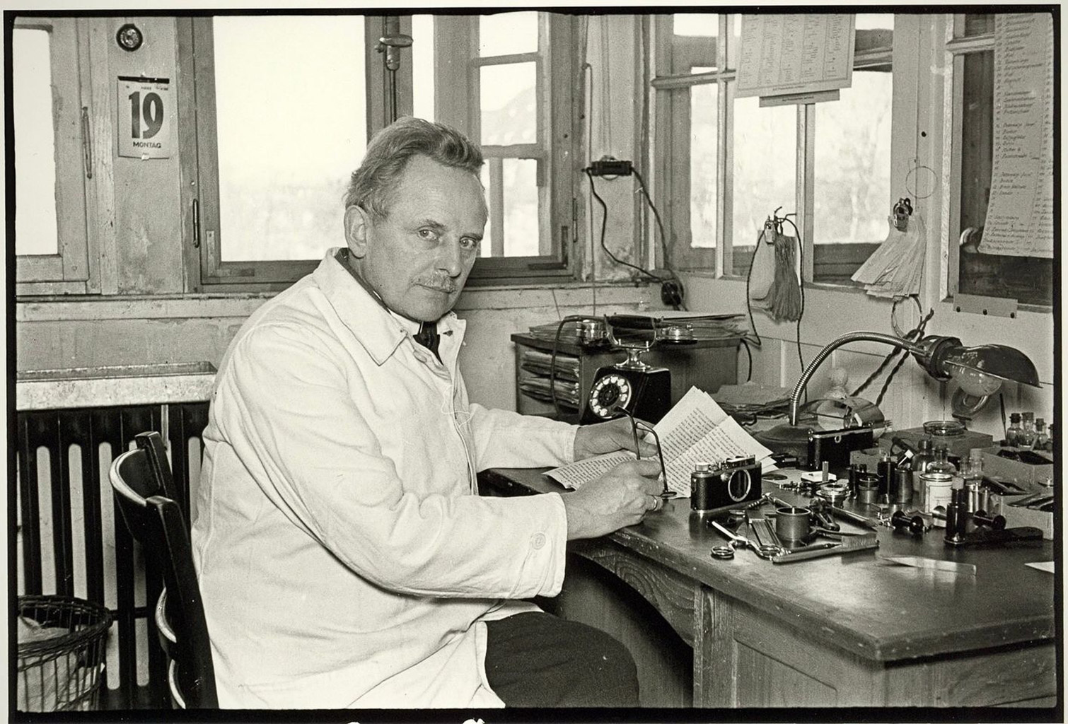 A man in a lab coat sits at a desk filled with scientific instruments, writing notes while looking directly at the camera. The room features large windows and a calendar hanging on the wall.