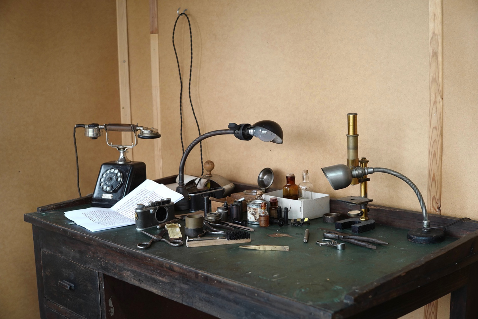 A vintage workspace featuring an old rotary telephone, various scientific instruments, glass containers, and paperwork on a dark wooden desk.