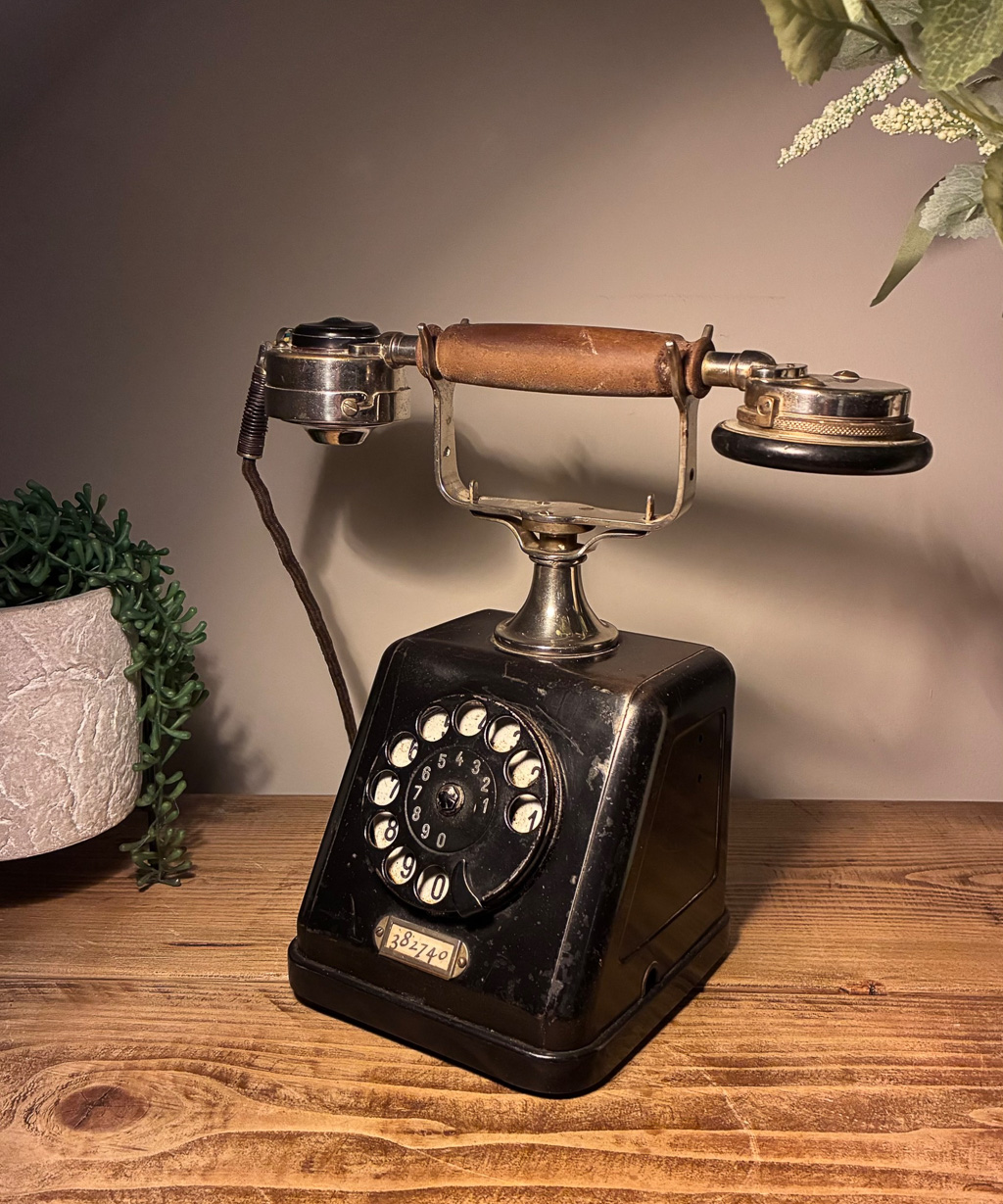 A vintage black rotary telephone with a wooden handle, displayed on a wooden table next to a potted plant.