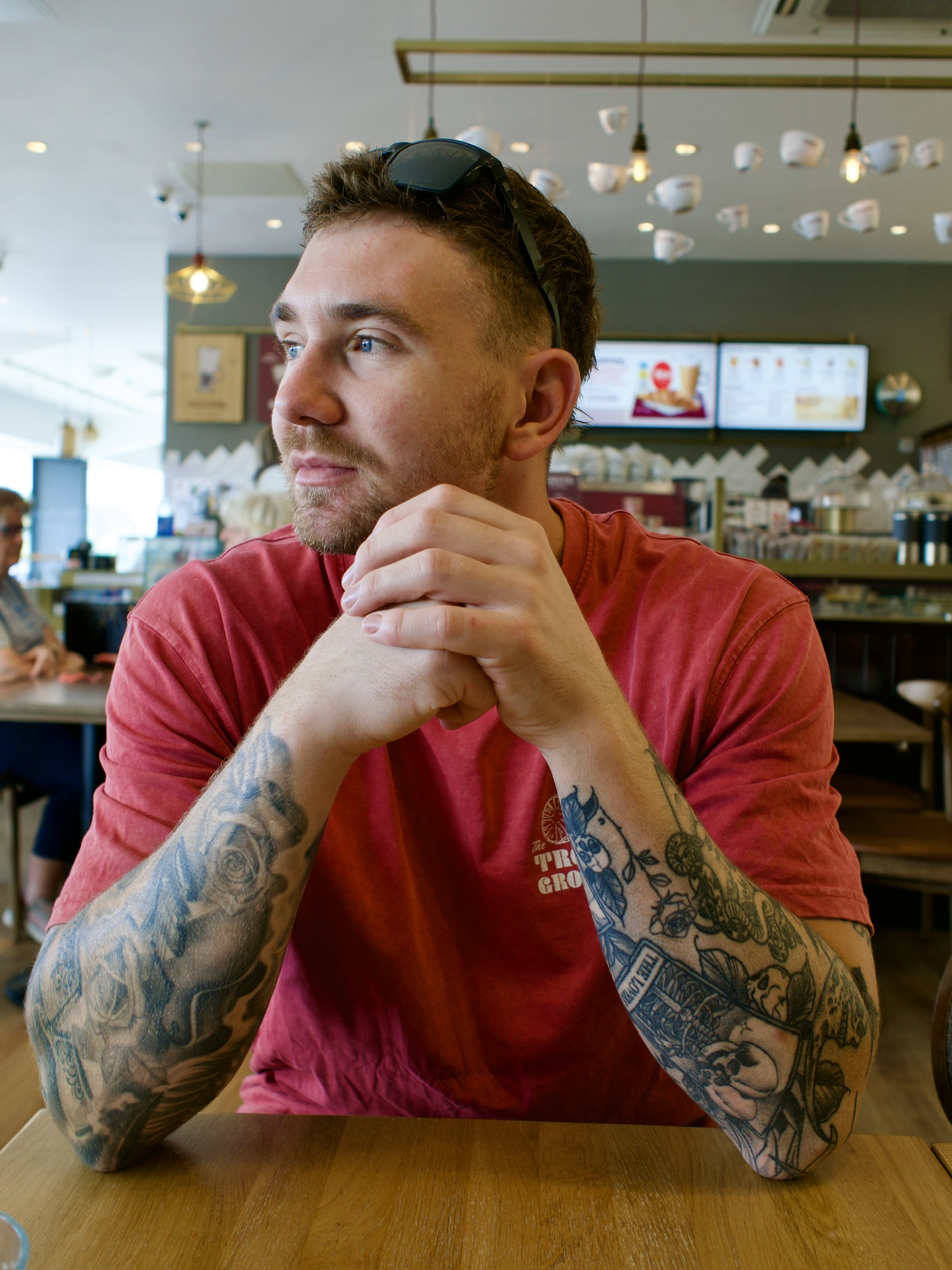 A man with heavily tattooed forearms rests his chin on his clasped hands at a cafe table, sunglasses pushed up on his head.