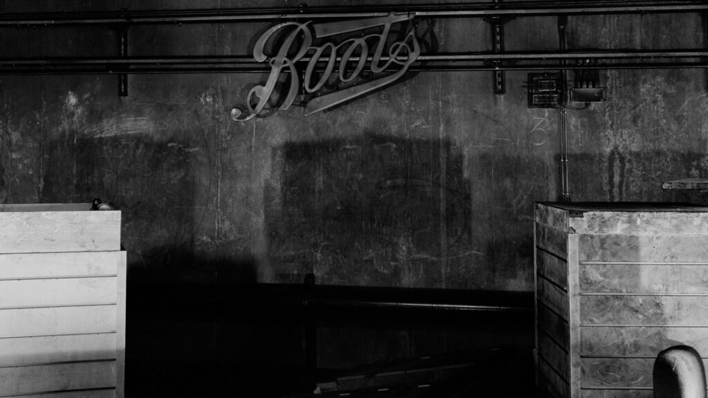 Black and white photograph of a wall with the word 'Boots' in stylized lettering, featuring two wooden crates in the foreground.
