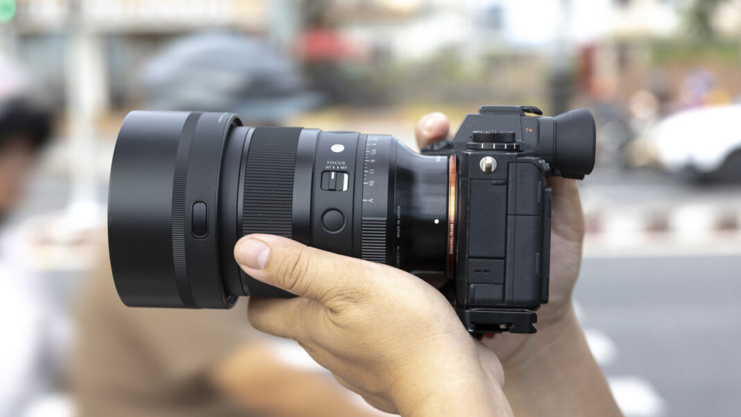 A photographer in a cap and glasses holding a mirrorless camera to his eye in a blurred street scene.