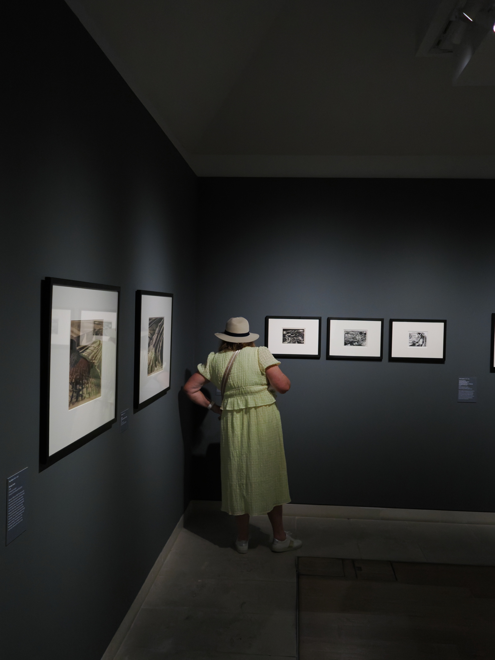 A visitor in a light green dress and hat observing framed artworks on a dark grey wall in an art gallery.