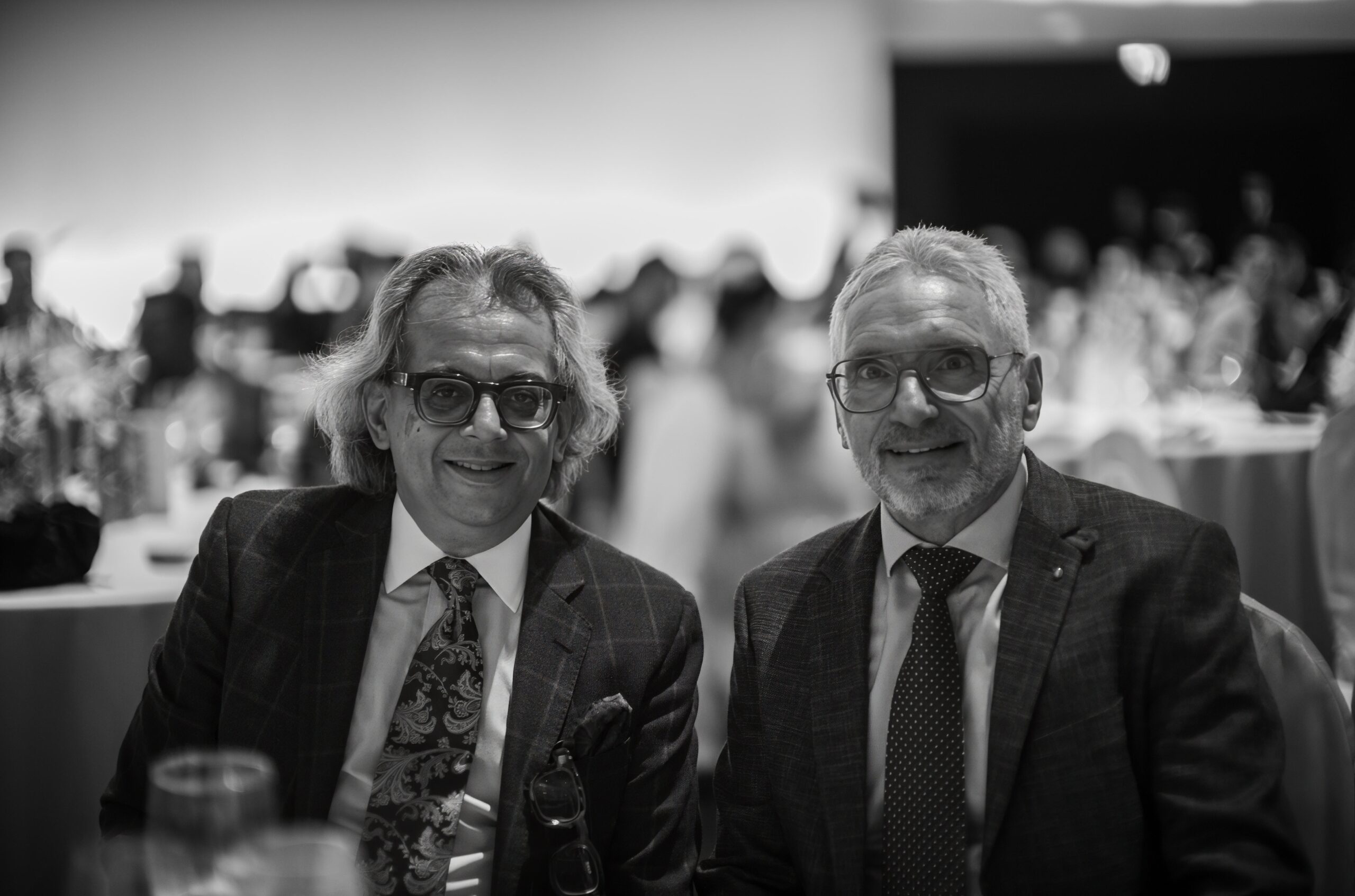 Two men smiling for the camera at a social event, dressed in suits. The background shows a blurred setting with other attendees.