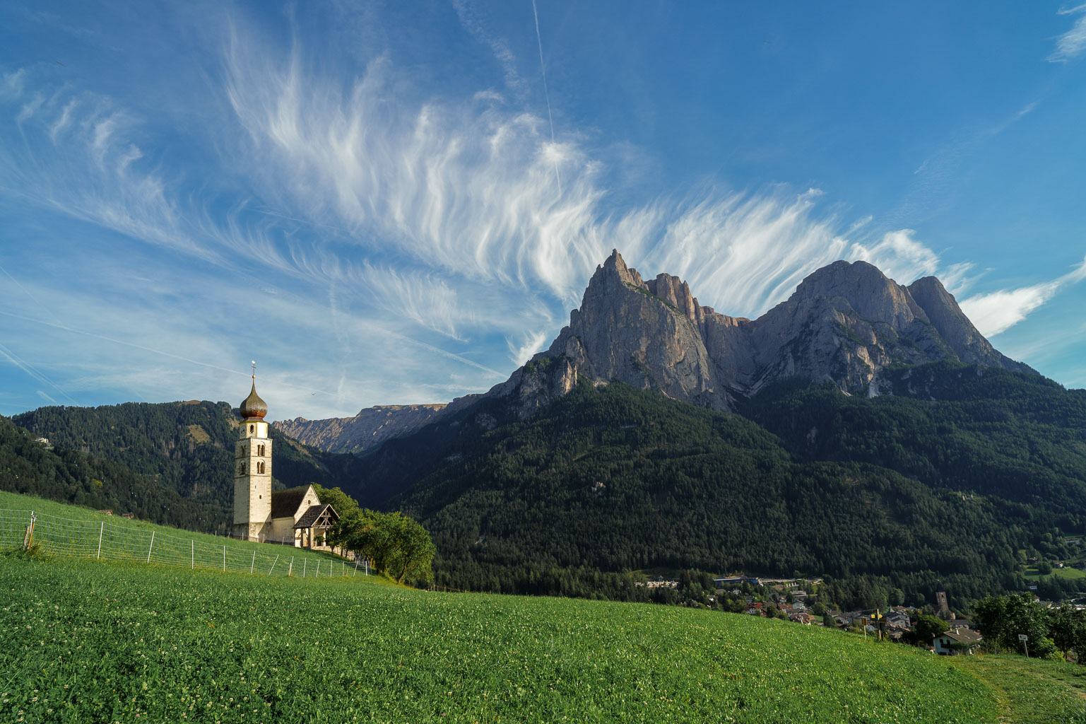 A scenic view featuring a church with a tall spire set against a backdrop of dramatic mountains and a clear blue sky, surrounded by green fields.