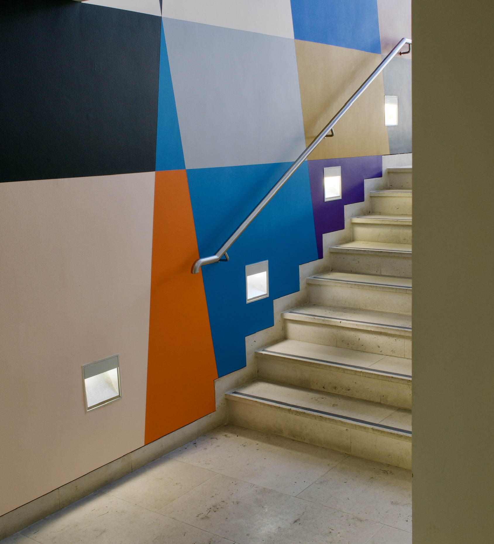 A stone staircase with a metal handrail set against a wall painted in geometric blocks of navy, orange, blue and cream.