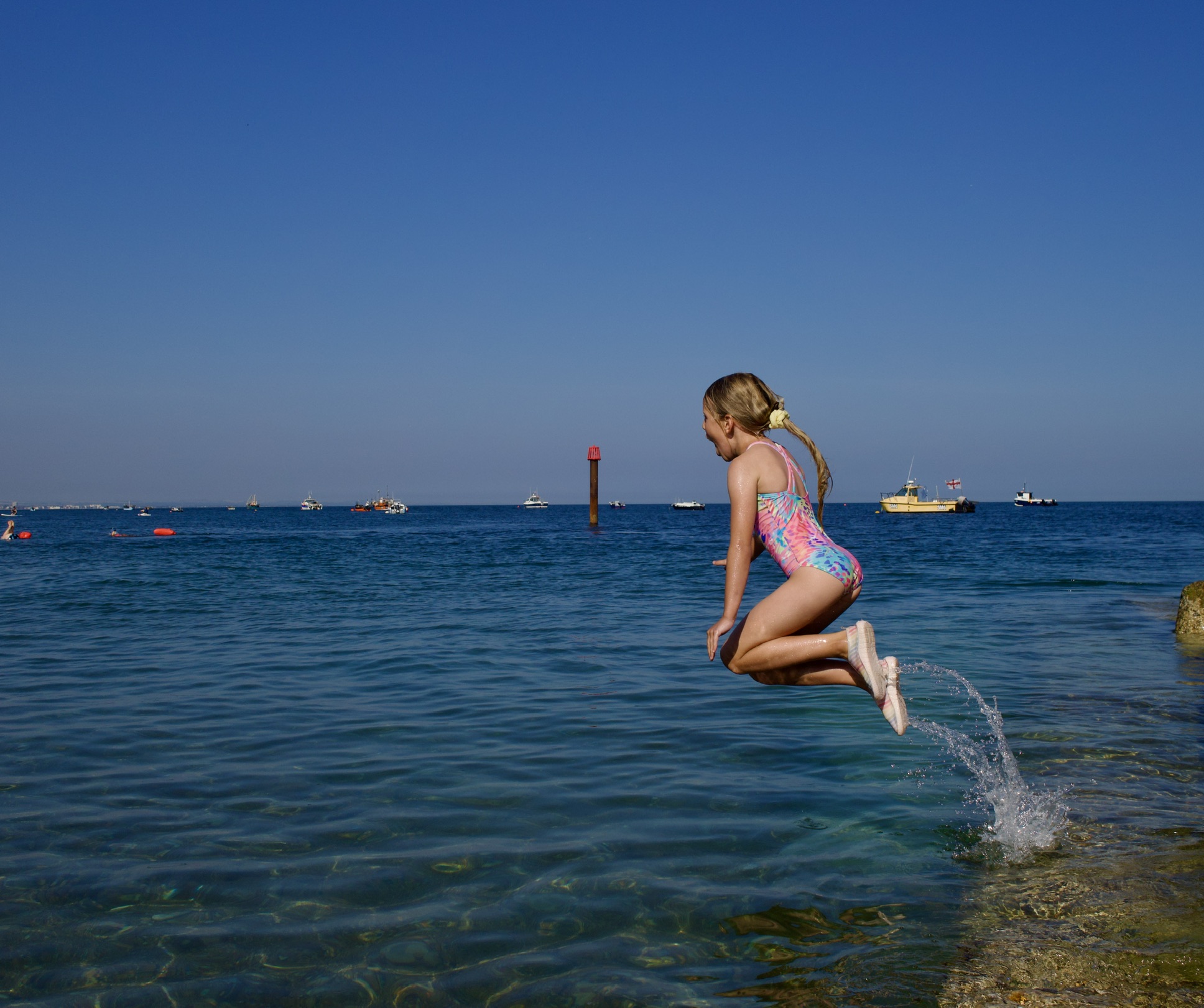 A girl in a colourful swimsuit leaps into the sea from a harbour wall, with fishing boats moored beyond.