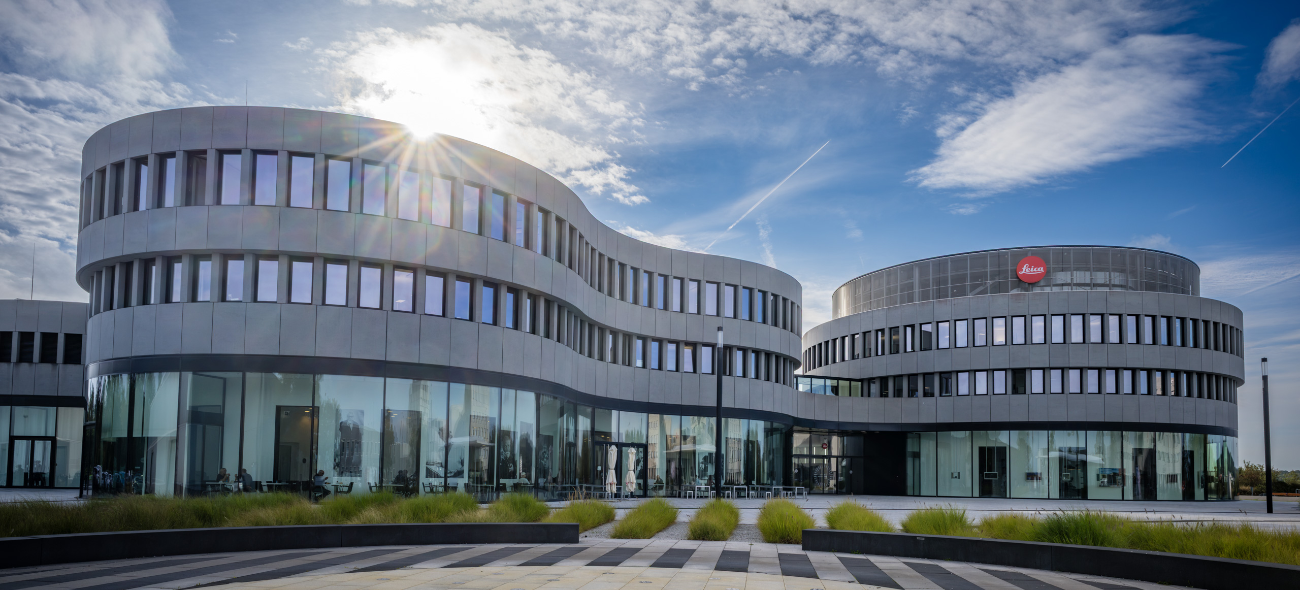 Leica Wetzlar visit: The curved glass-and-concrete Leica Campus building in Wetzlar, lit by low sun, with the red Leica logo on its facade.
