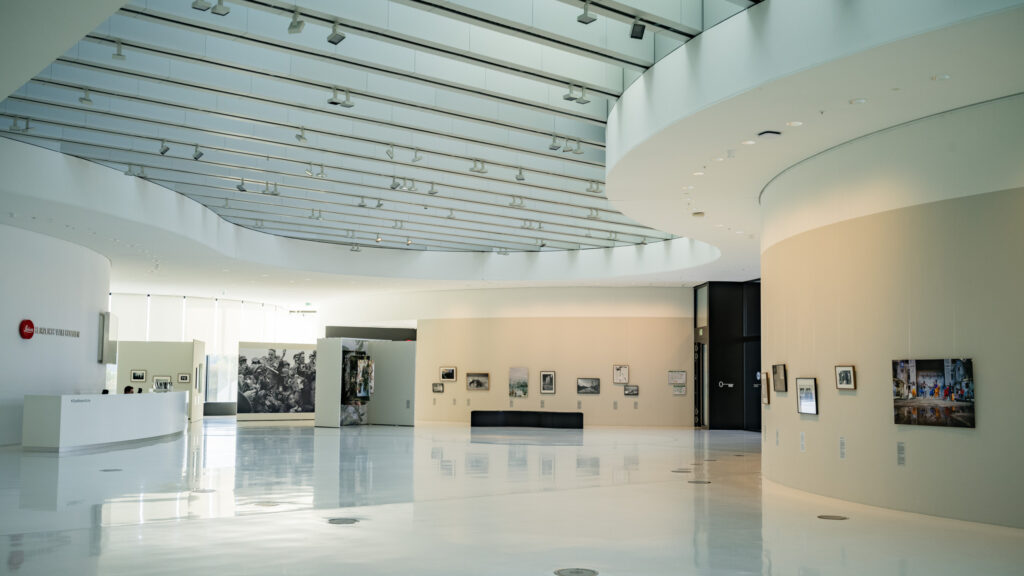 Leica Wetzlar visit: A bright museum entrance hall with a reception desk, a bench, and rows of small framed photographs on a curved wall.