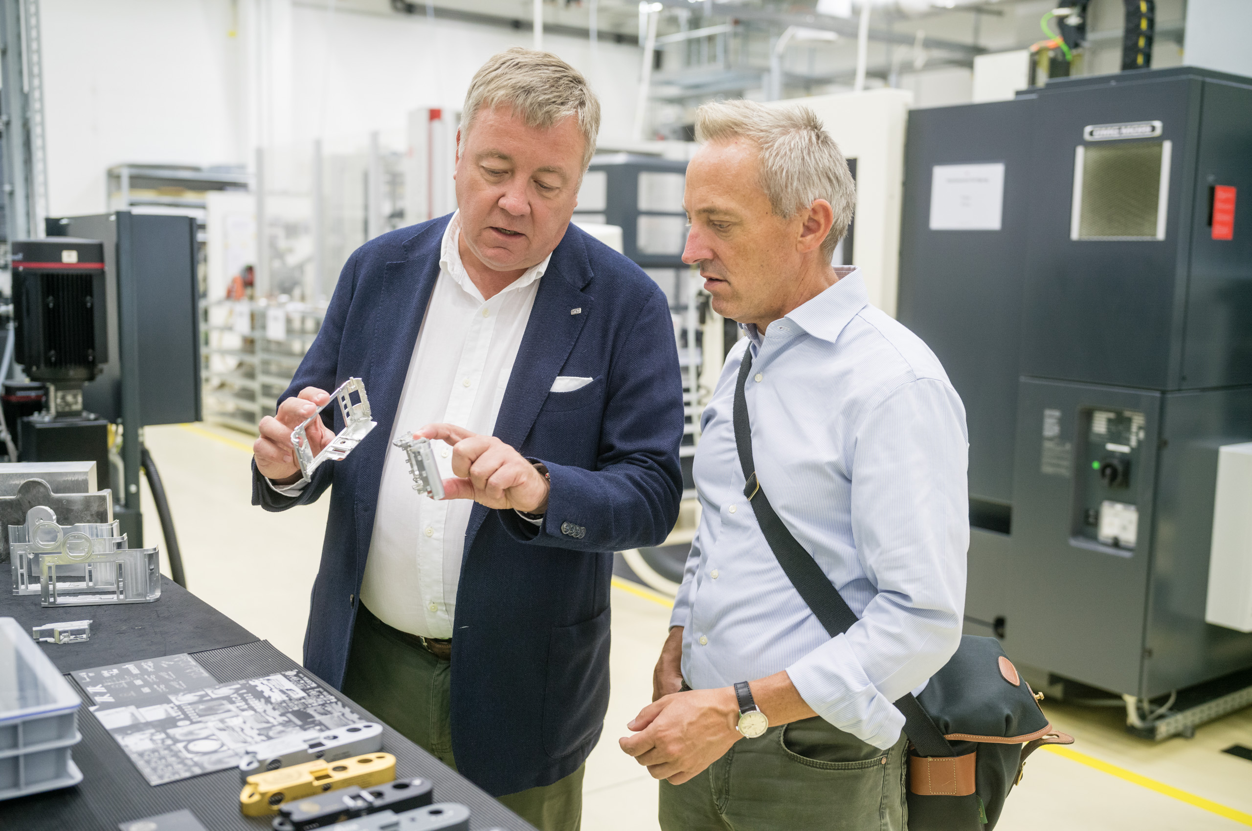 Leica Wetzlar visit: Two men in a factory workshop examining small metal camera-body components on a workbench, with high-tech machinery behind them.