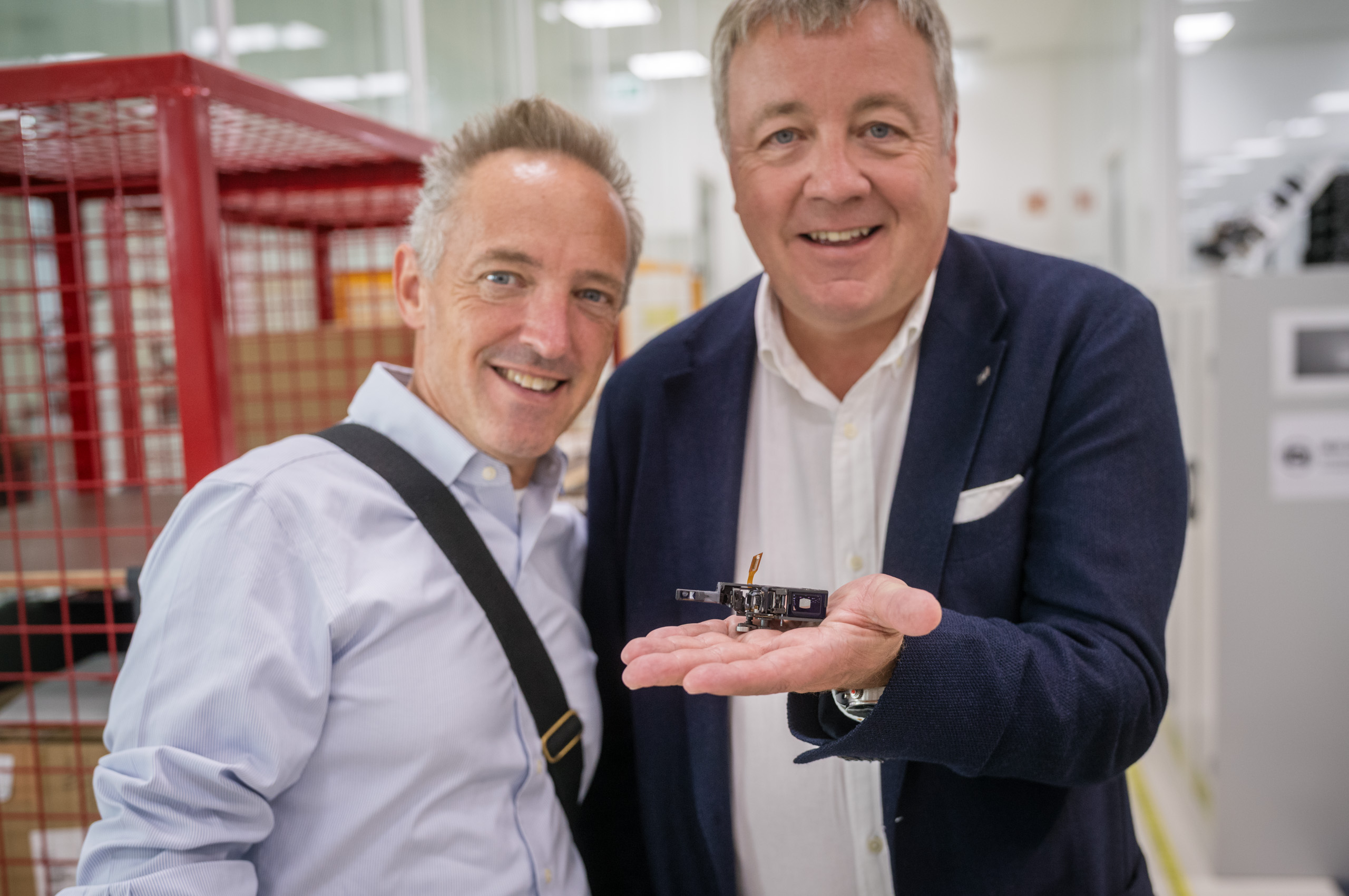Leica Wetzlar visit: Two smiling men in a factory cleanroom, one holding a tiny camera component (rangefinder) in his open palm.