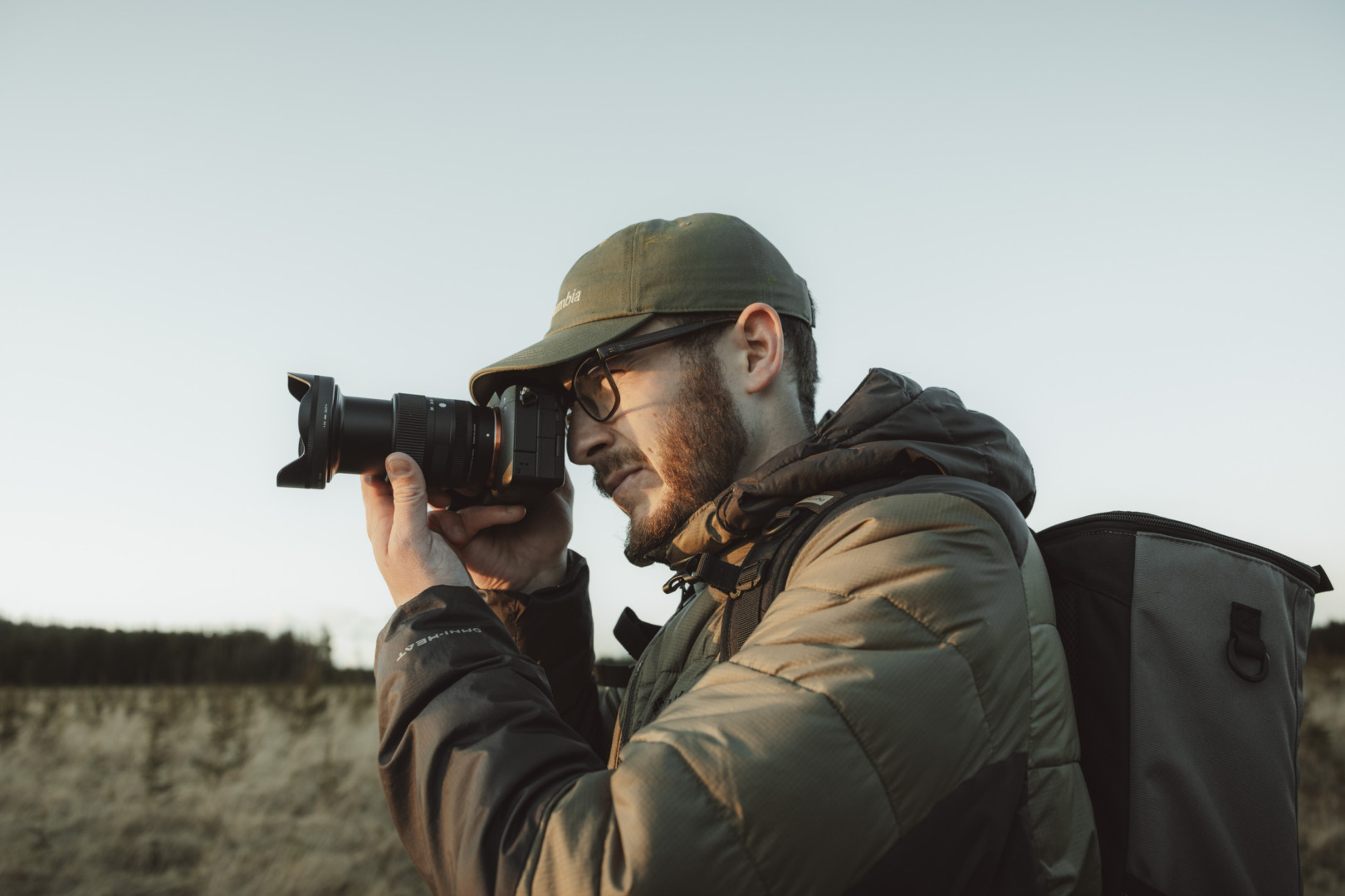 A photographer in a cap and glasses holding a mirrorless camera to his eye in a blurred street scene.