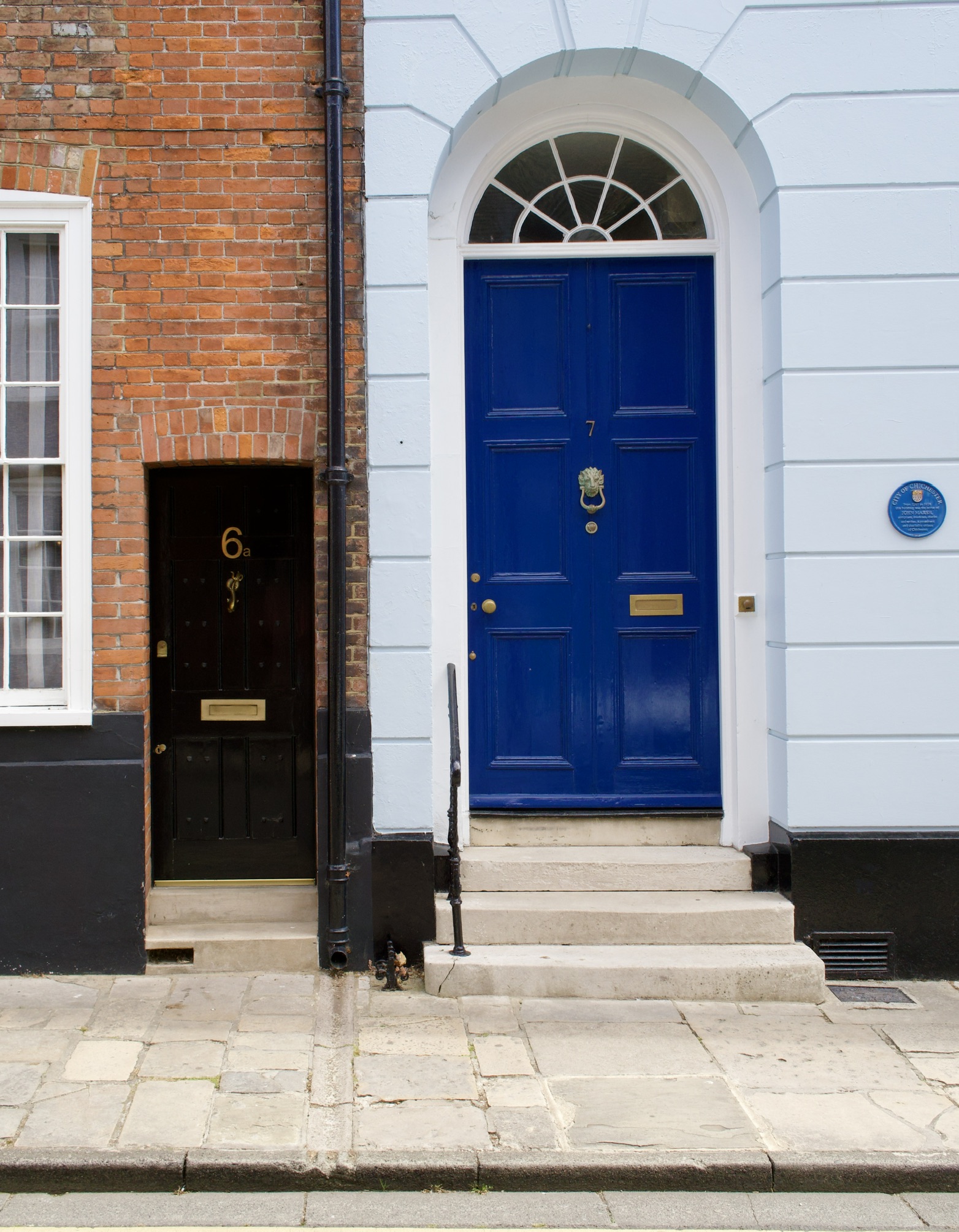A small black front door numbered 6a beside a taller blue Georgian front door numbered 7, in a brick and painted terrace.
