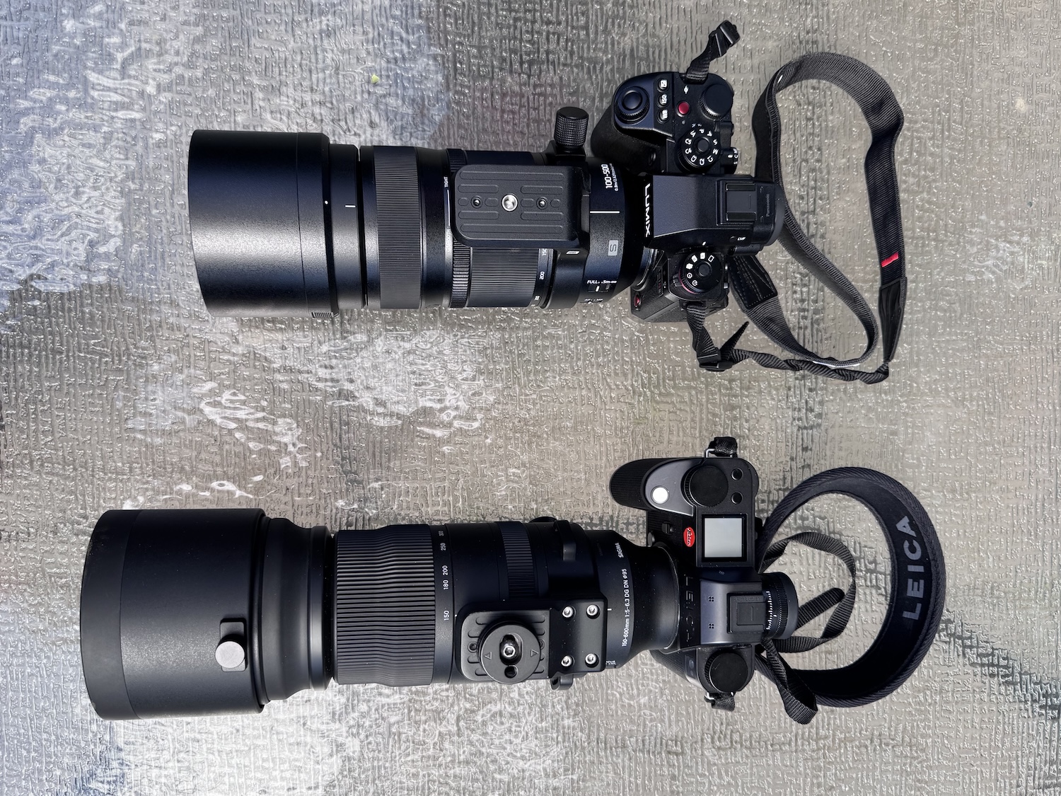 A Lumix S1R Ii with a telephoto zoom above a Leica SL3-S with a telephoto zoom on a glass table top.