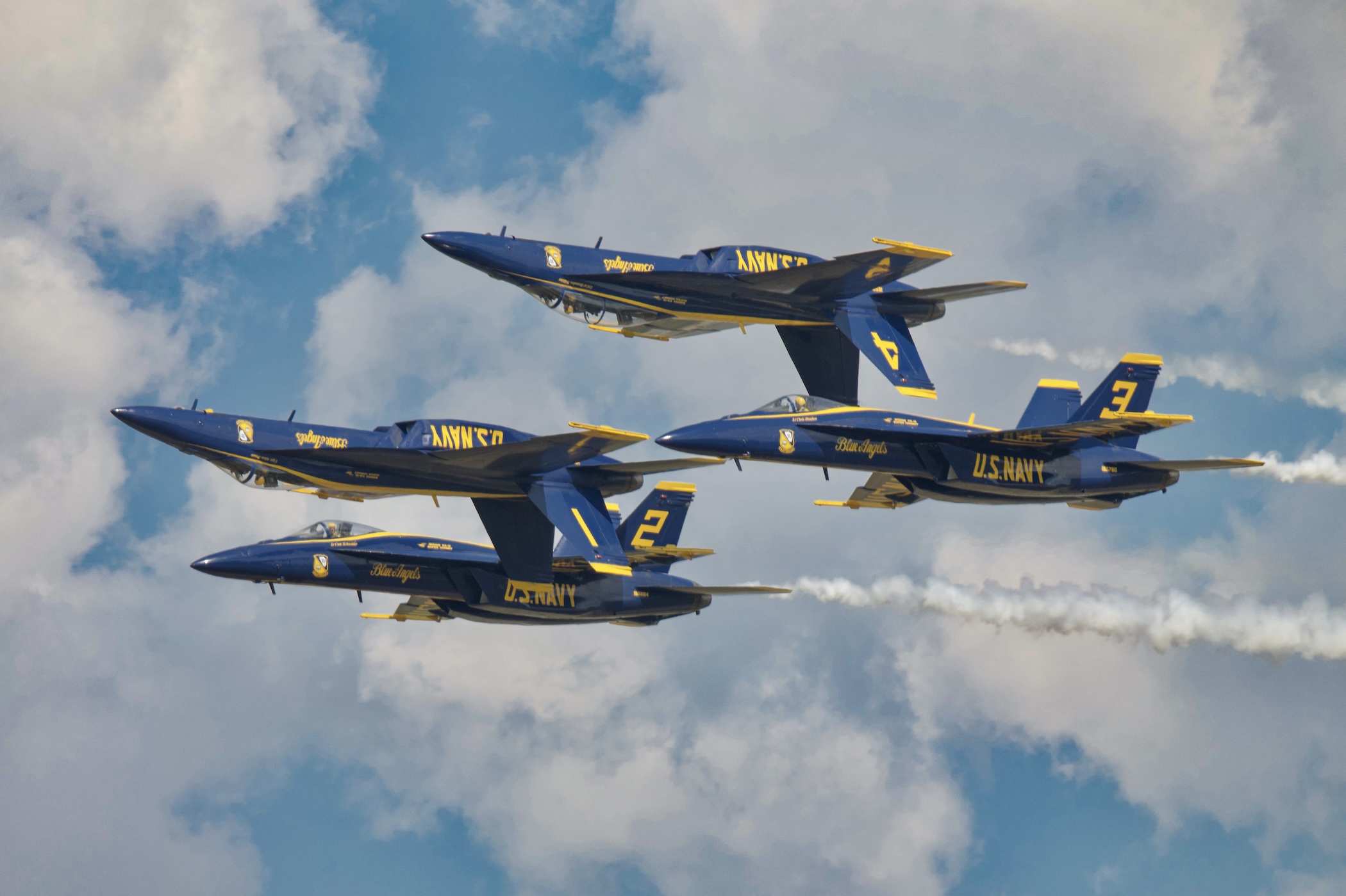Four Blue Angels F/A-18s in diamond formation banking together, smoke trailing from two of the jets.