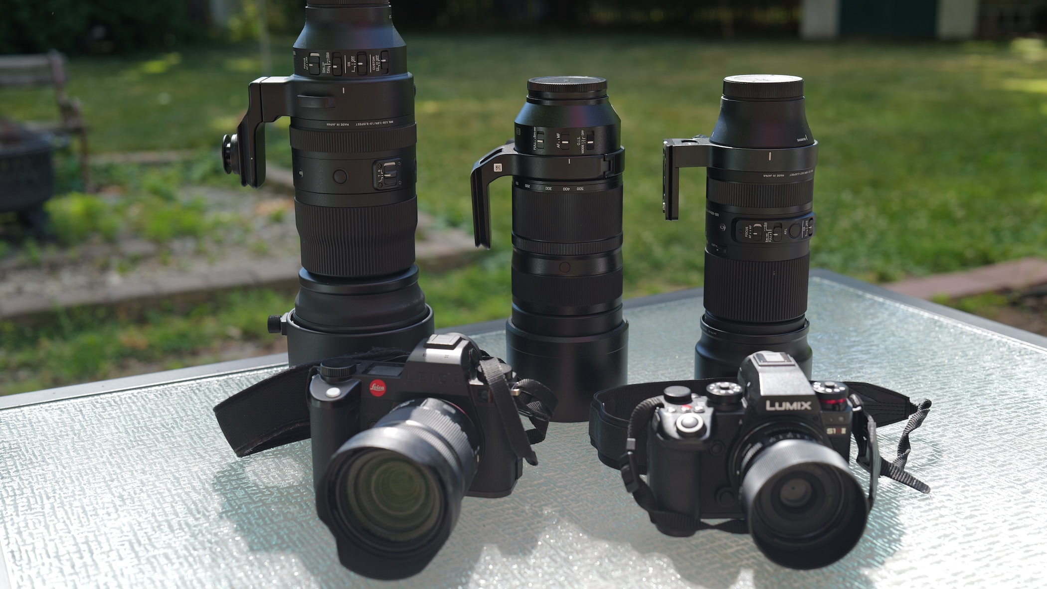 Three telephoto zoom lenses and two mirrorless cameras, a Leica and a Panasonic Lumix S1, arranged on a glass patio table with a lawn behind.