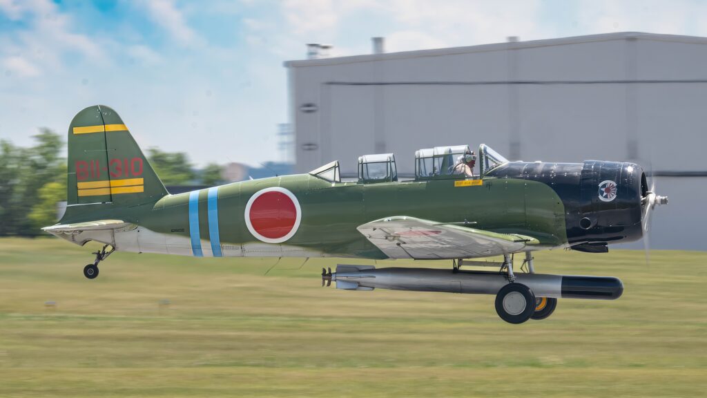 A green and blue Japanese Kate torpedo bomber replica warbird with red roundels taking off in front of a hangar.