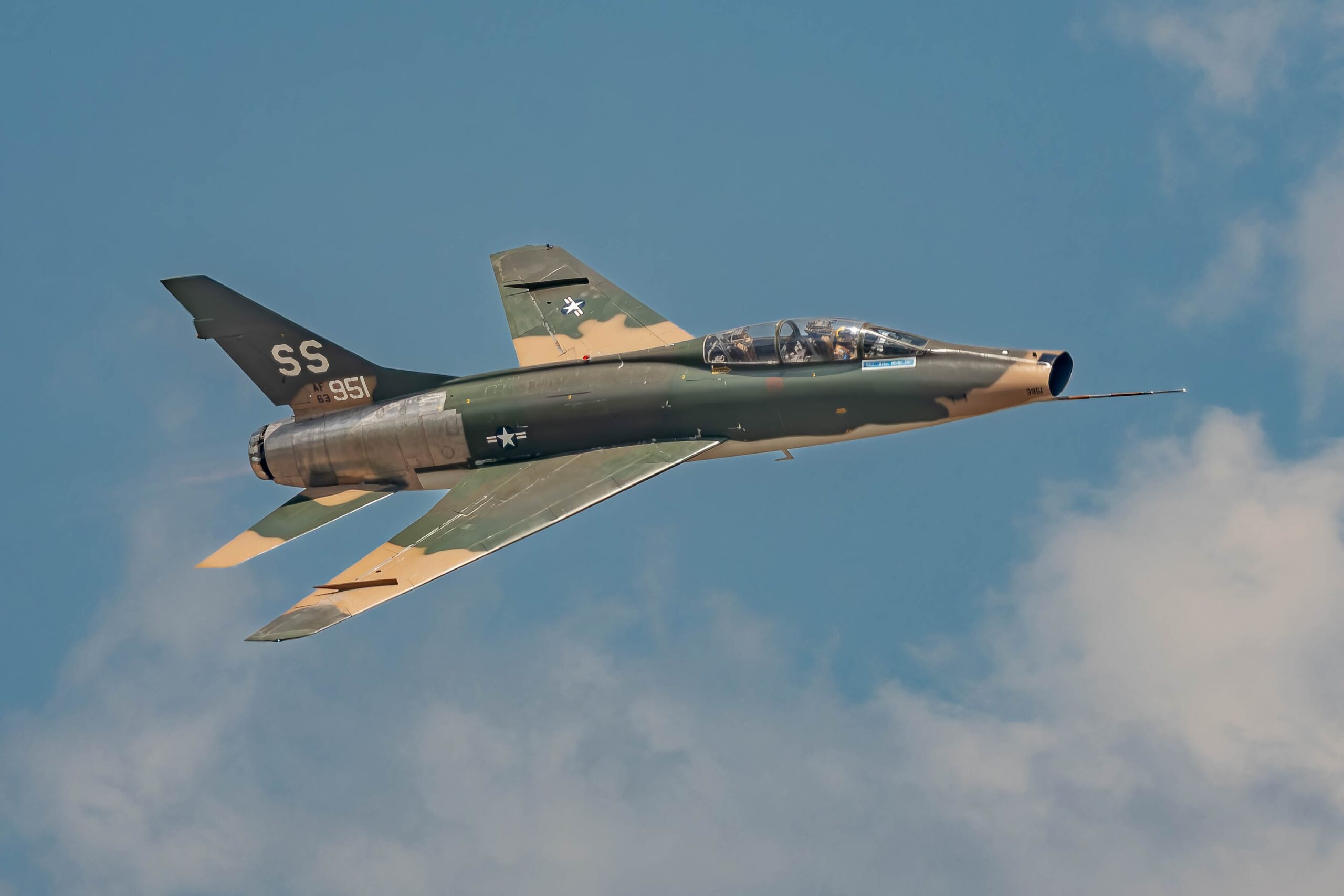 A camouflaged North American F-100 Super Sabre banking against a blue sky with a long trailing pitot boom.