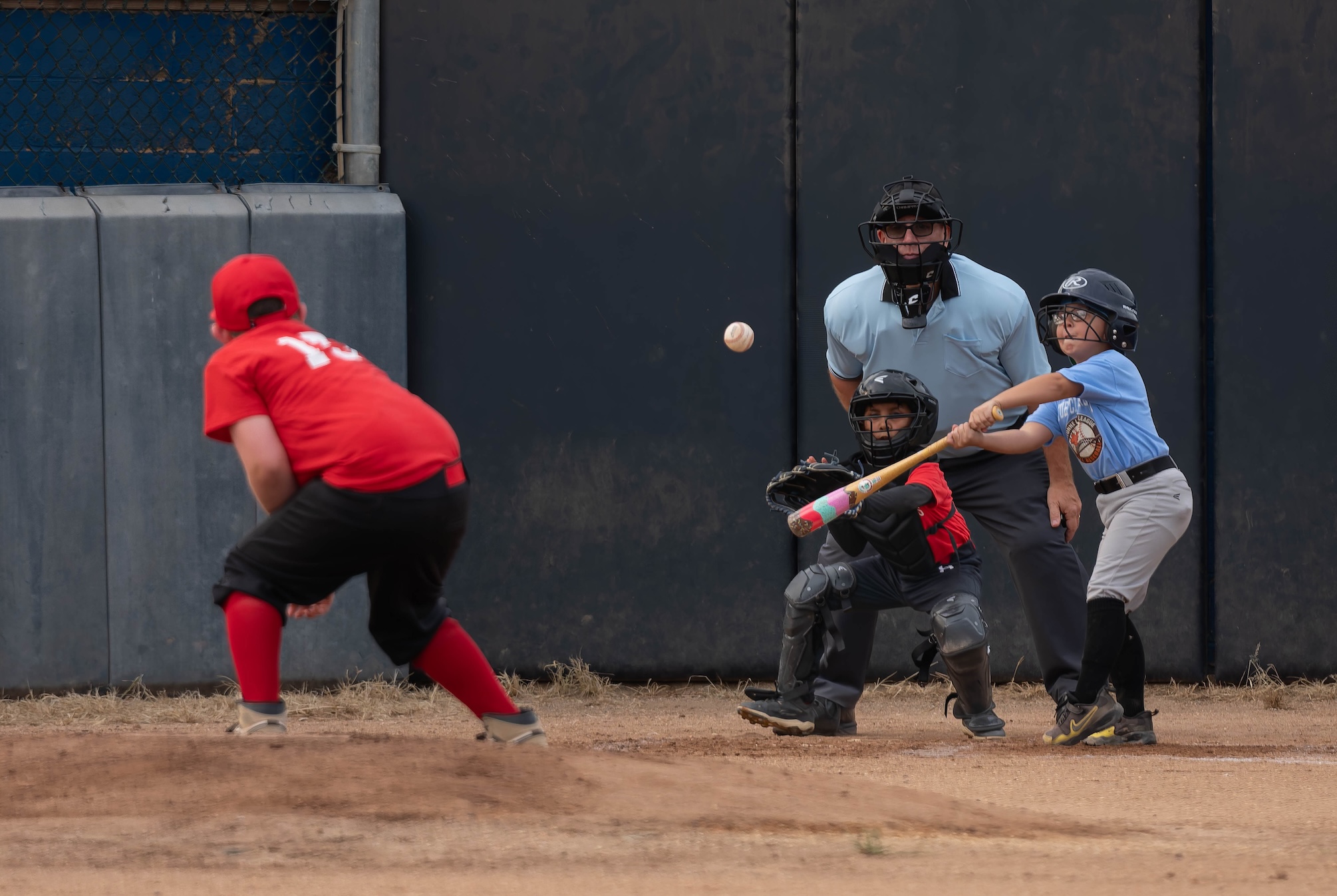 A young batter connecting with a pitch as the catcher and umpire watch behind home plate.