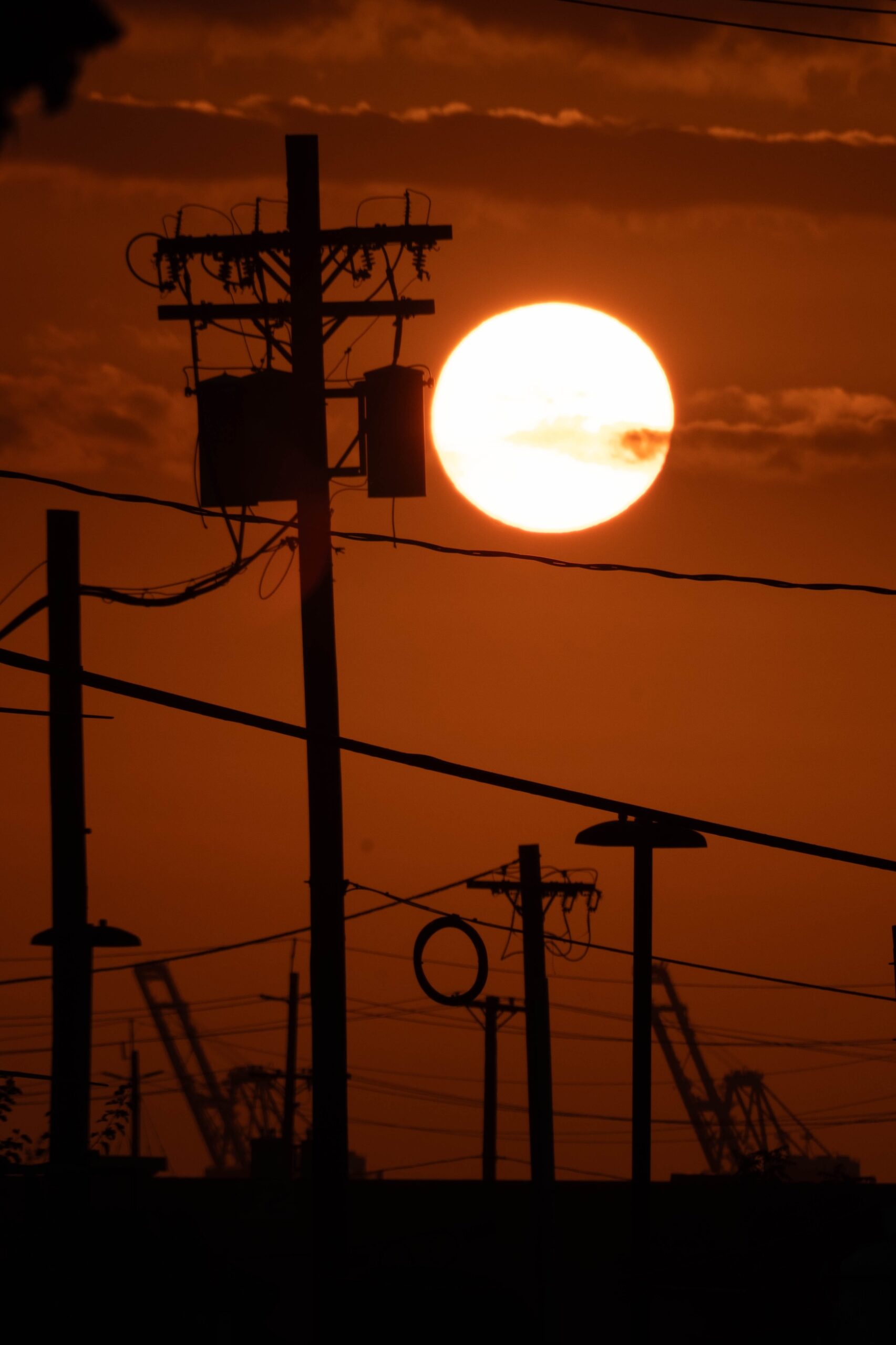 The setting sun glowing behind silhouetted utility poles and dockside cranes at dusk.