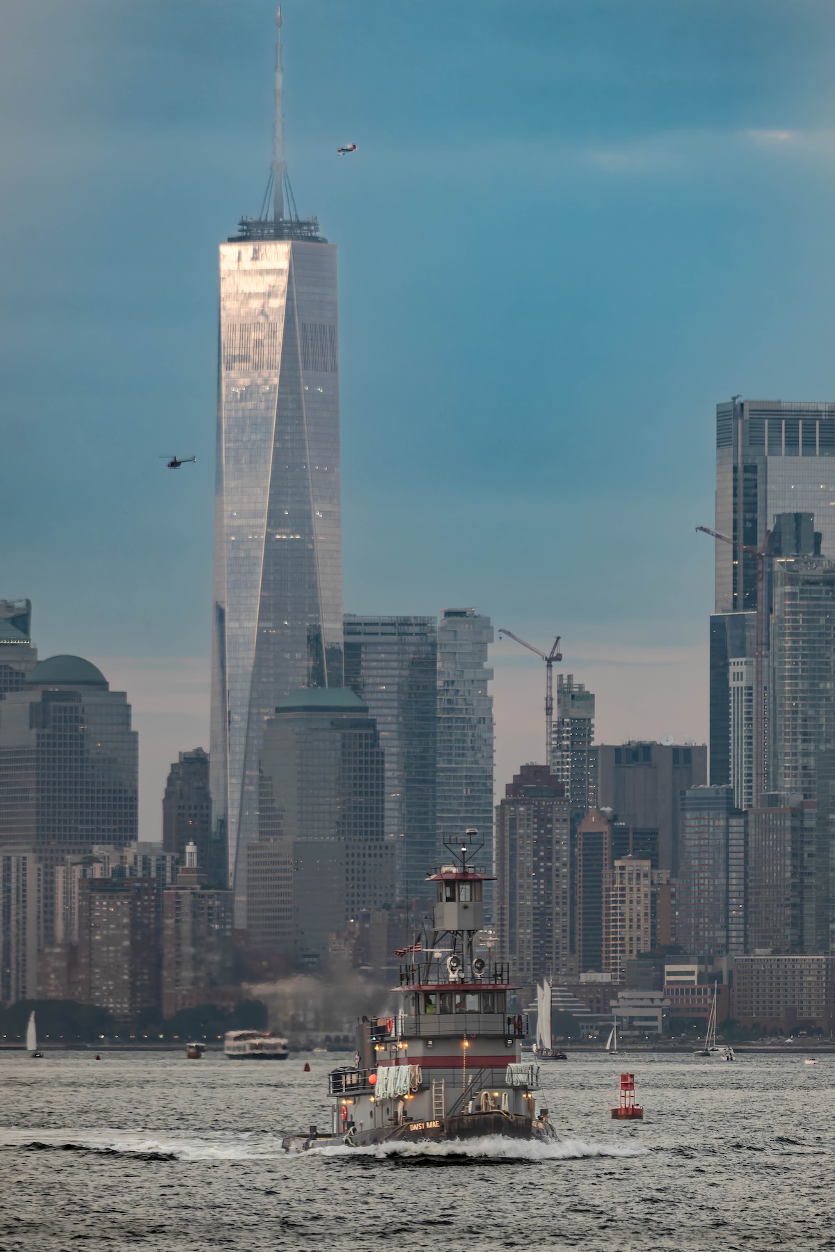 One World Trade Center catching the evening light above the Hudson River, with helicopters overhead and a tugboat crossing in front.